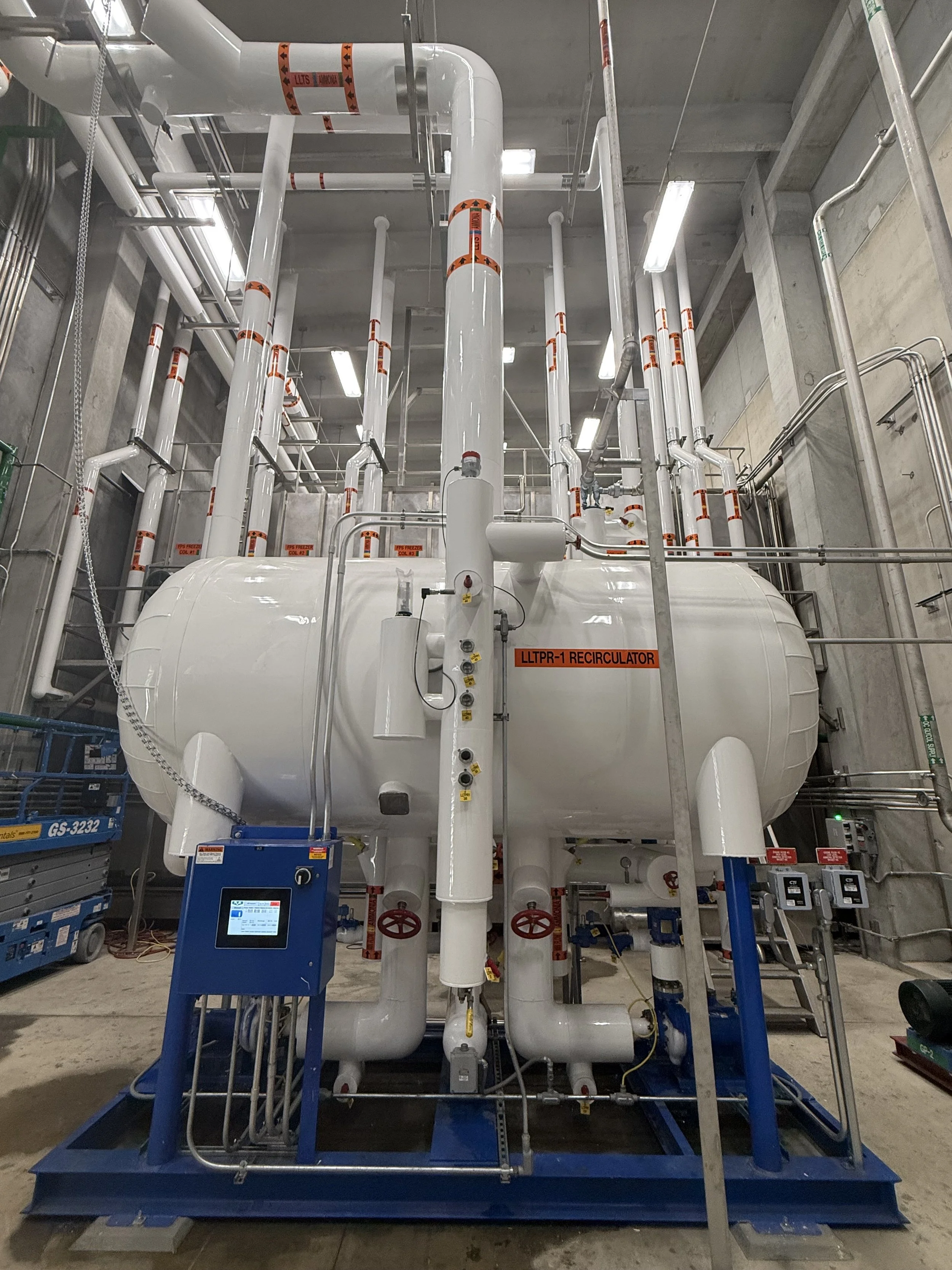 Industrial water or gas tank with pipes and control panels in a construction site or facility. The tank is labeled 'LLTPR-1 Recirculator'.