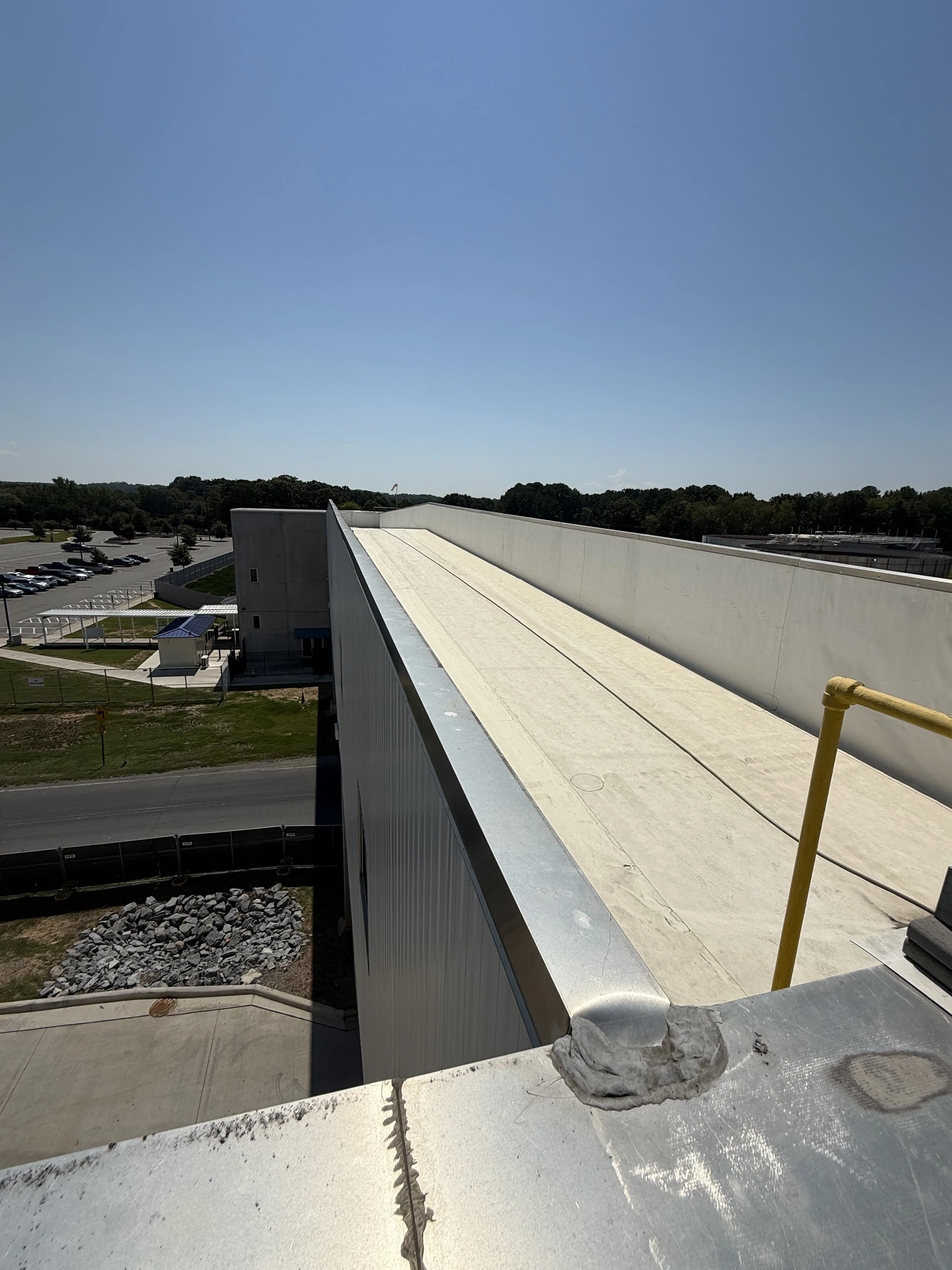 Construction site view from a high vantage point showing a large flat rooftop, part of a building with metal siding, parking lot with cars, trees, and a clear blue sky.