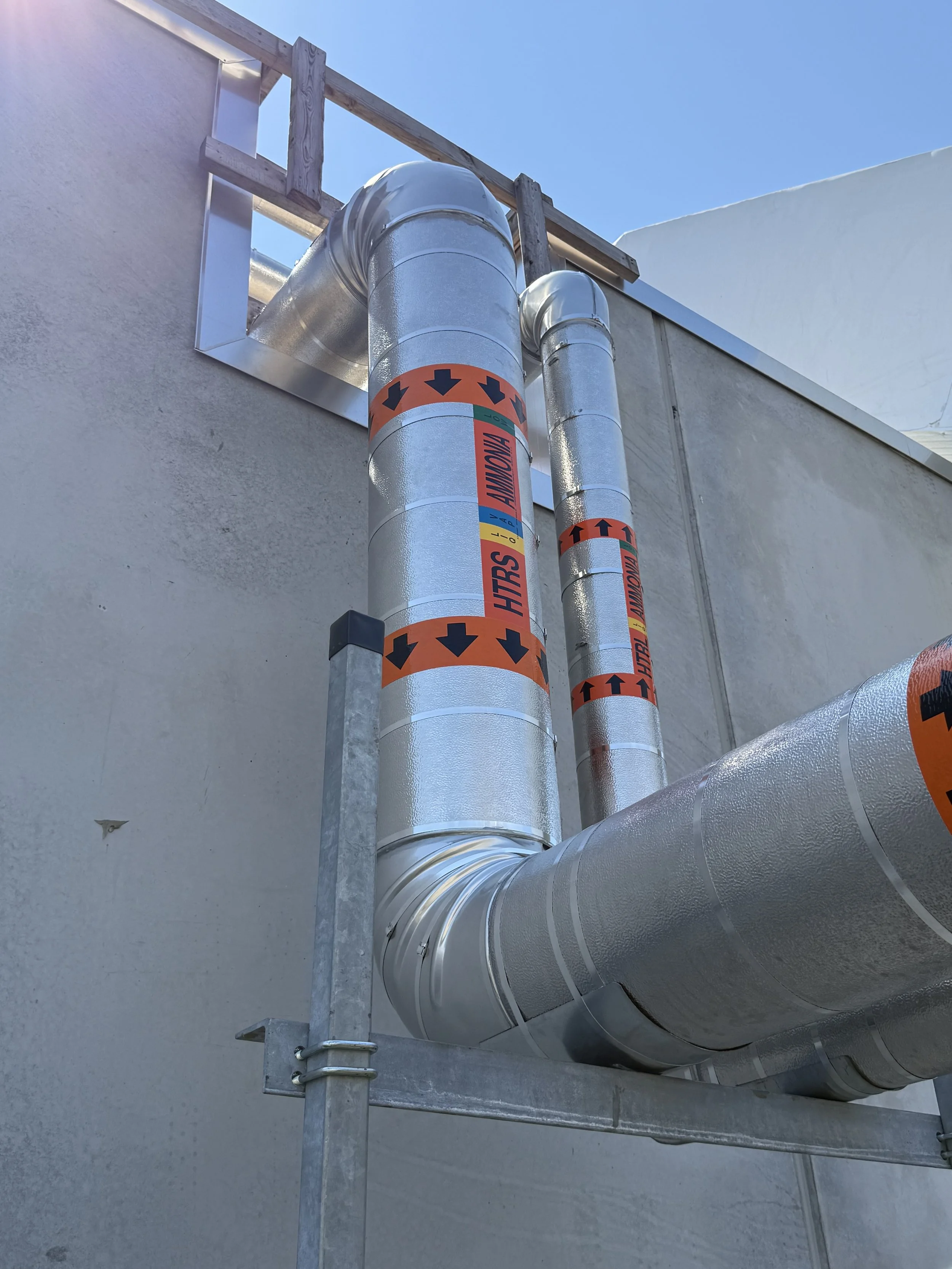 Silver insulated industrial pipes mounted along exterior wall of a building with an open sky above.