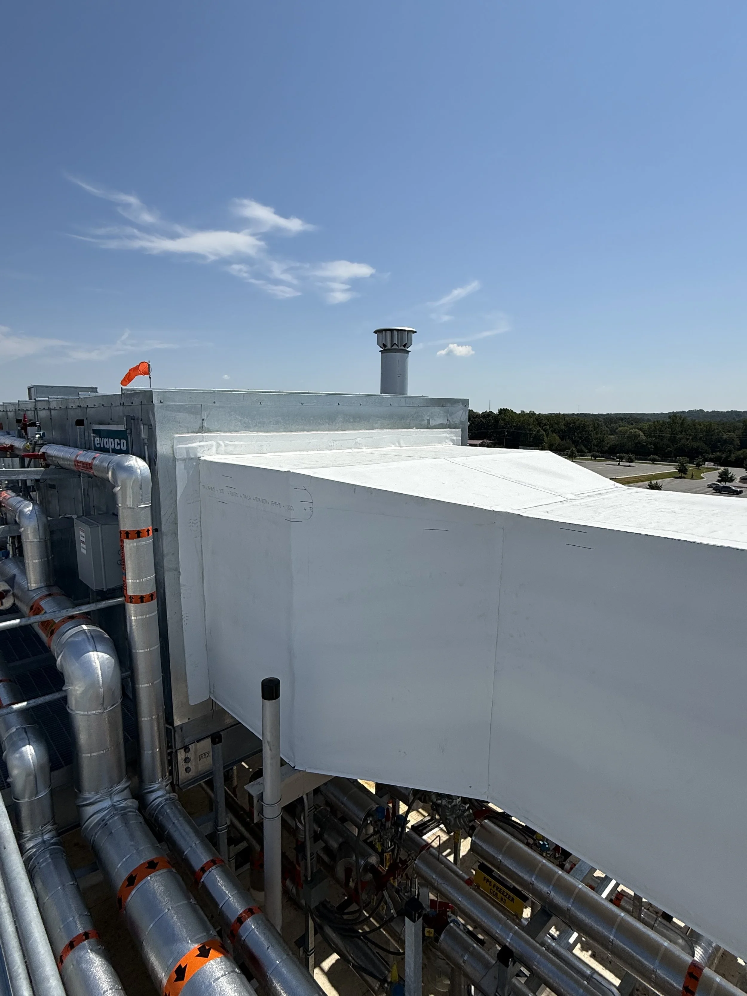 View of a rooftop with multiple metallic ventilation pipes and ducts, a vent pipe, and an air conditioning unit, with a background of trees and a clear blue sky with some clouds.