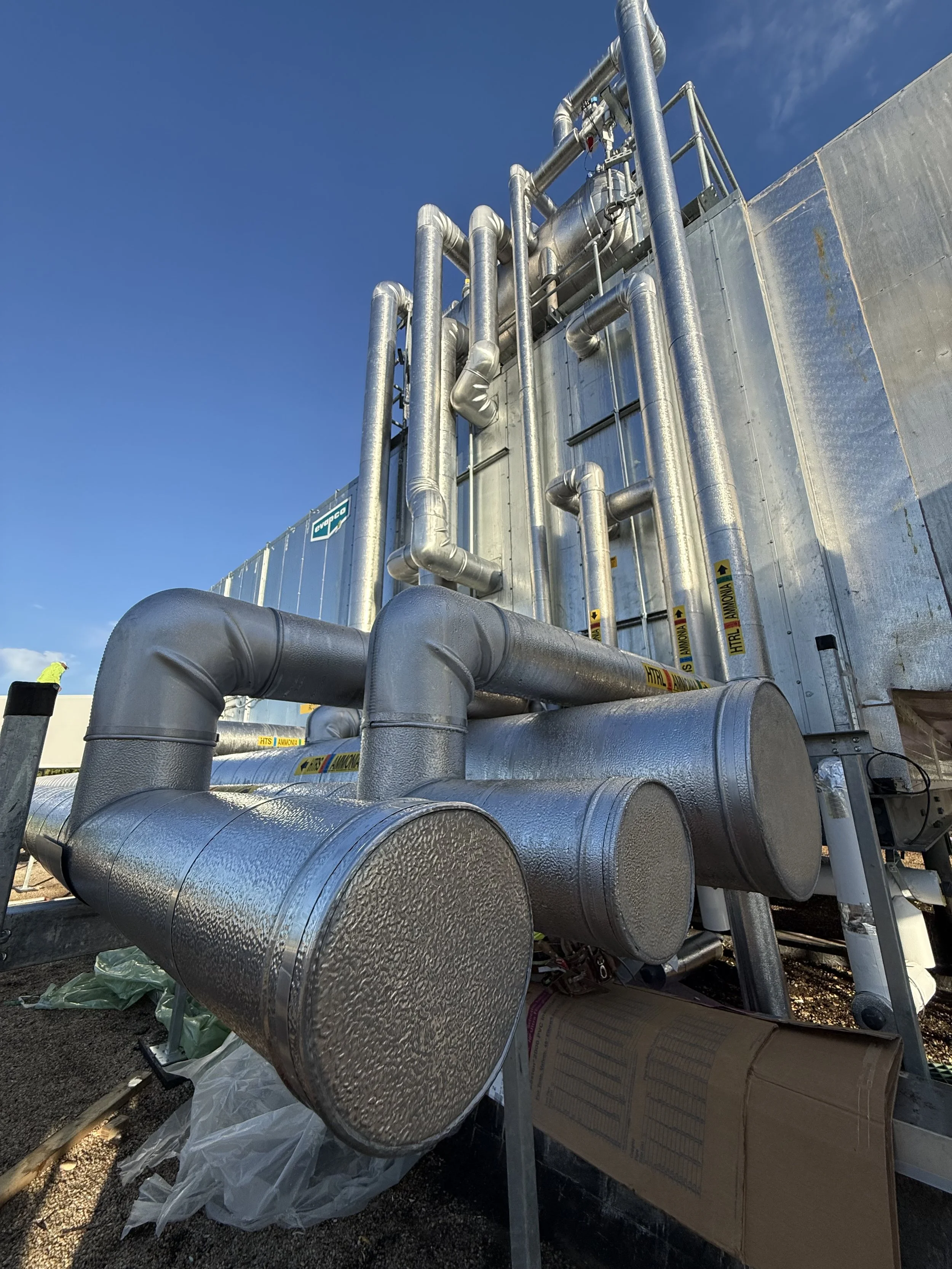 Industrial piping system with large metallic insulated pipes on a building exterior against a blue sky.