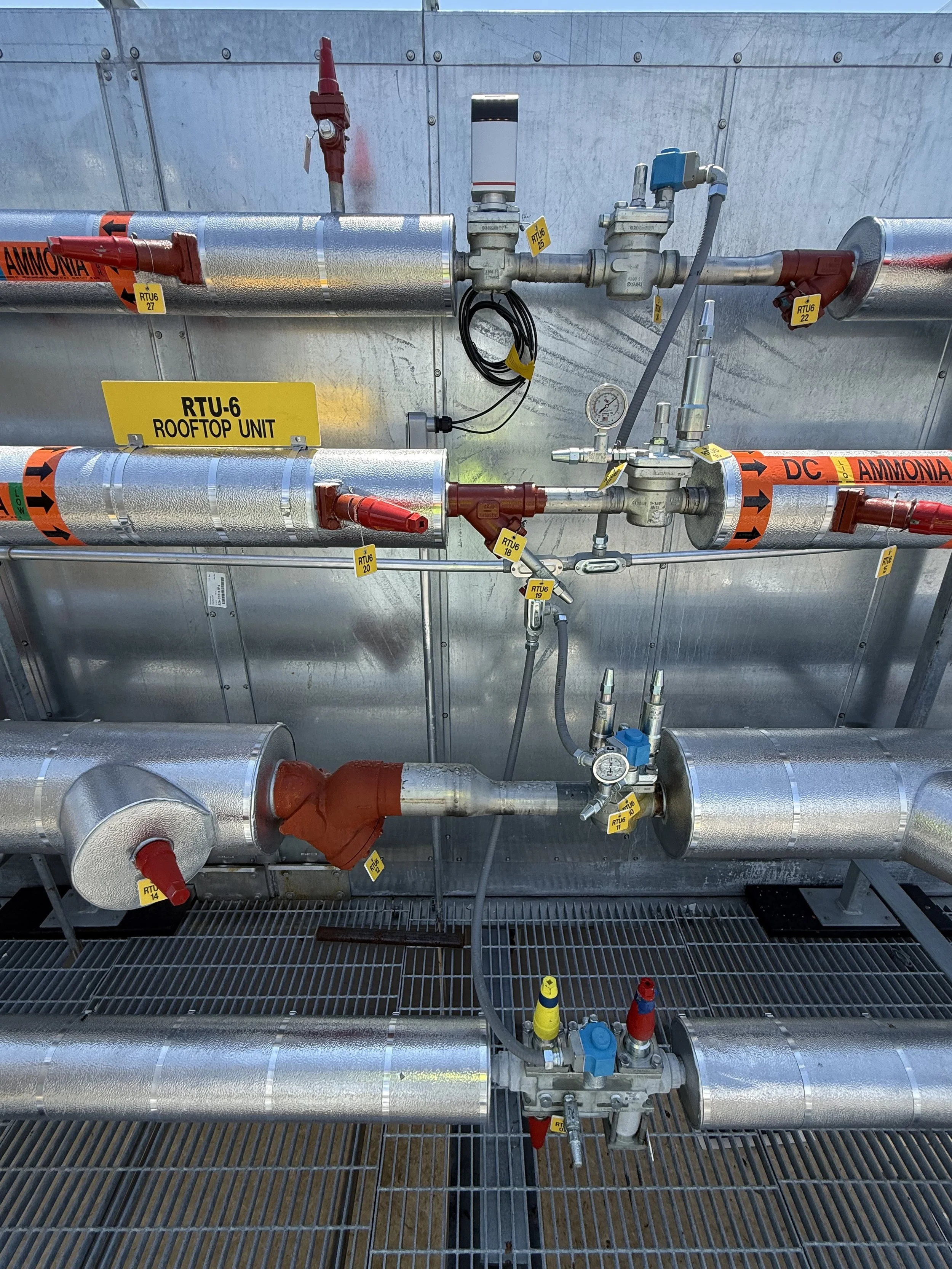 Industrial rooftop piping system with metal pipes, valves, gauges, and labels for ammonia and other gases, mounted on a grated platform.