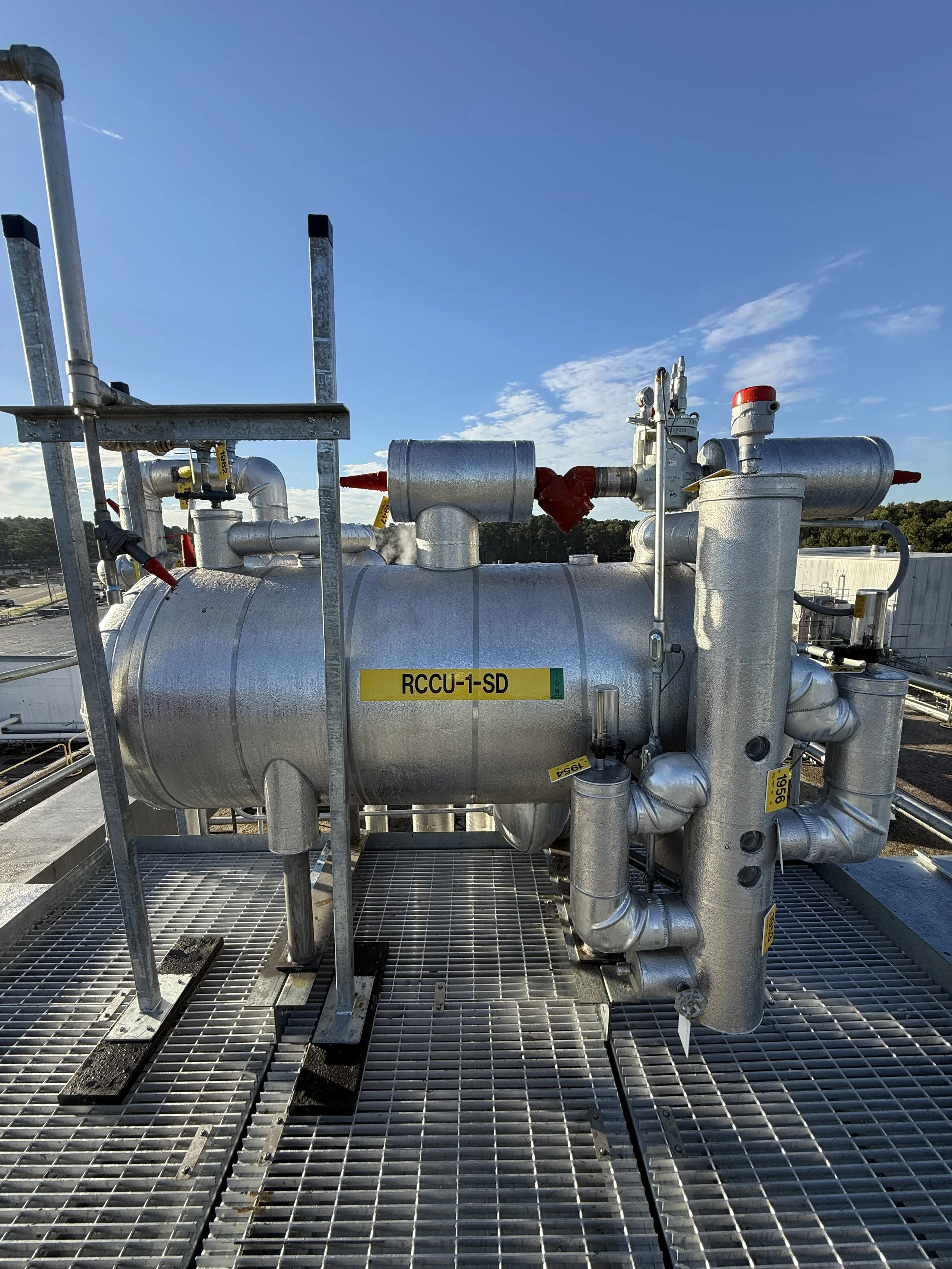 Industrial equipment with silver pipes and gauges on a metal grate platform outdoors against a blue sky with some clouds.