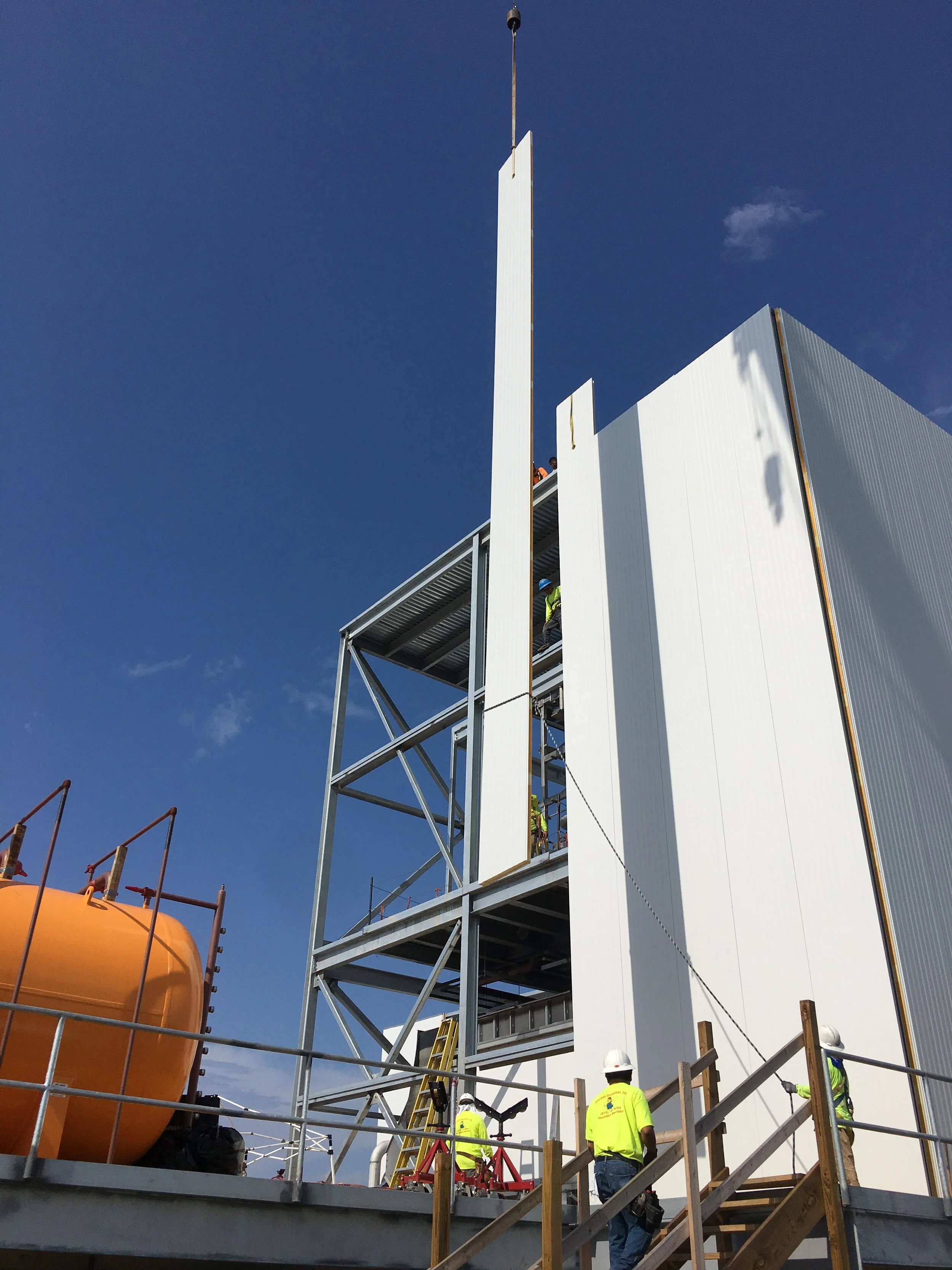 Construction workers in safety gear working on a building under construction with a blue sky background, a large orange tank visible on the left, and scaffolding and stairs.