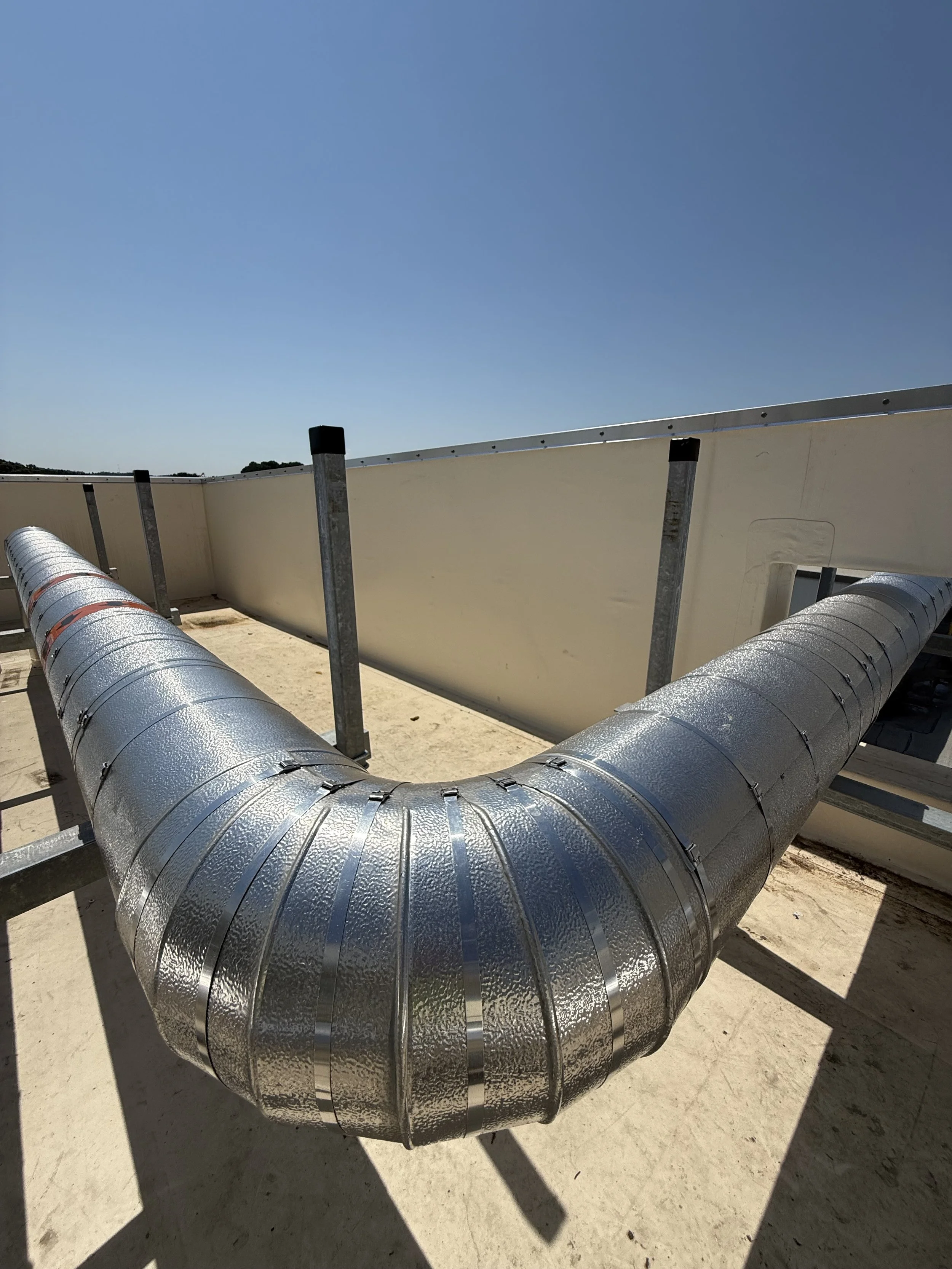 Large silver metallic pipes connected to an insulated ductwork system on a flat rooftop under a clear blue sky.