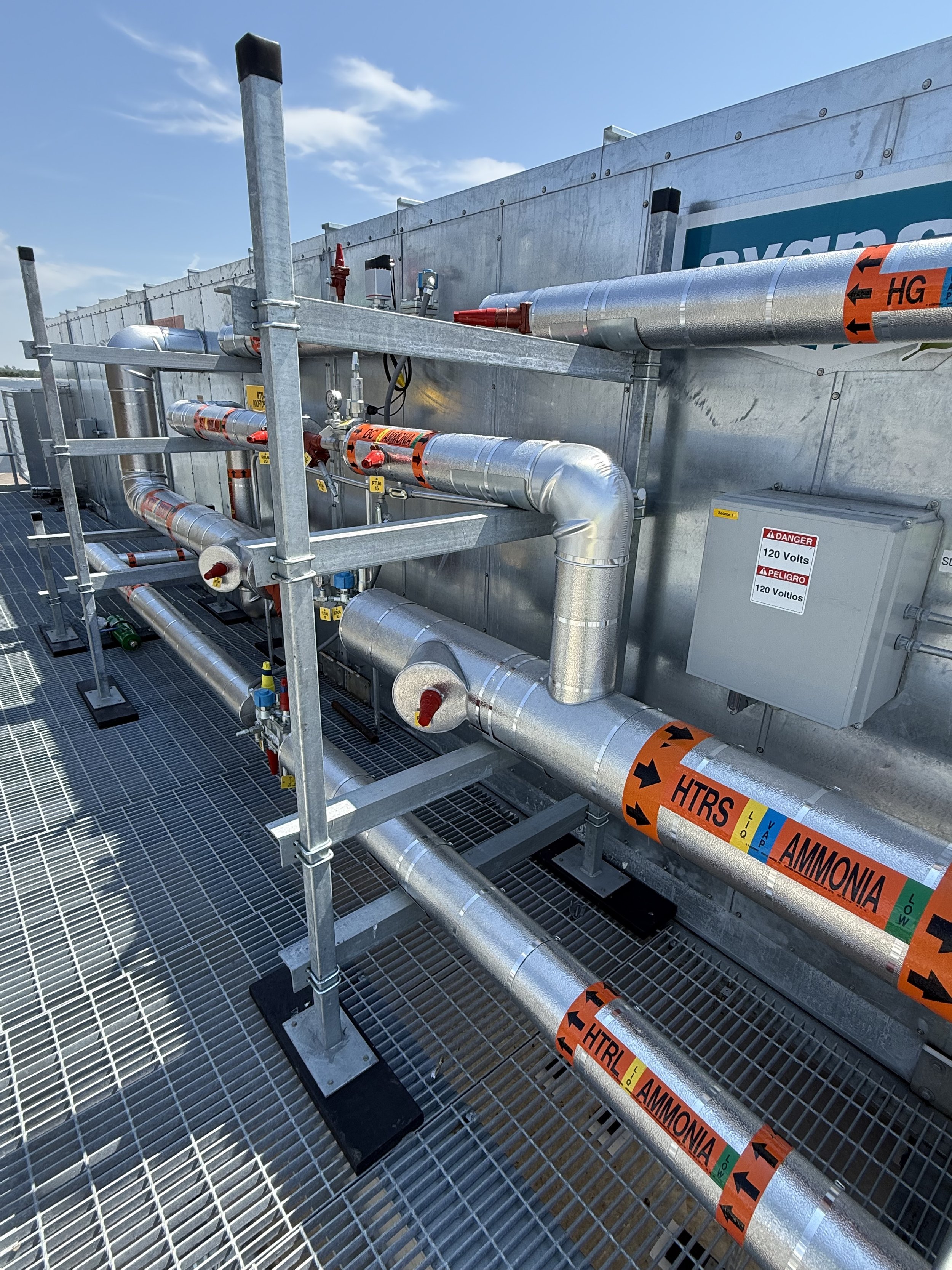 Industrial pipes and valves on a metallic rooftop with HVAC equipment, safety labels, and a grated metal surface, under a blue sky with some clouds.