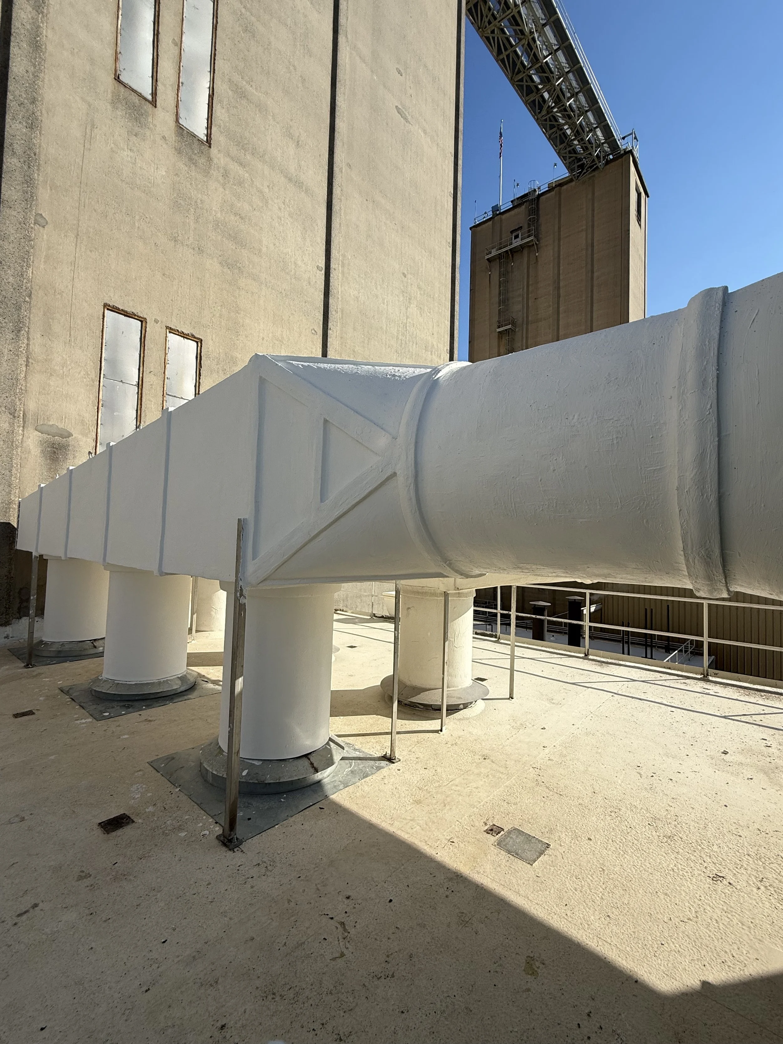 Industrial rooftop with large white ventilation ducts, a brick building with an American flag, and a metal bridge or walkway above.