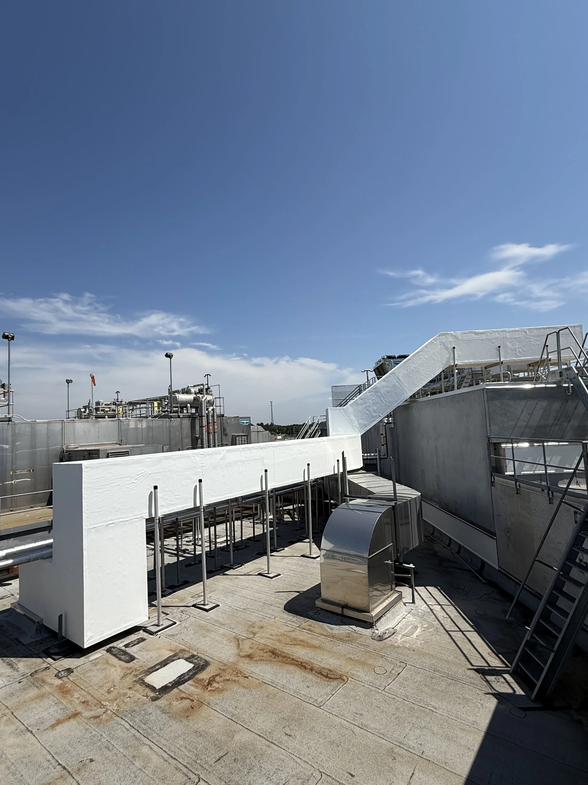 On a rooftop, there are industrial or mechanical structures made of metal, including stairs, vents, and pipes, with a clear blue sky overhead.