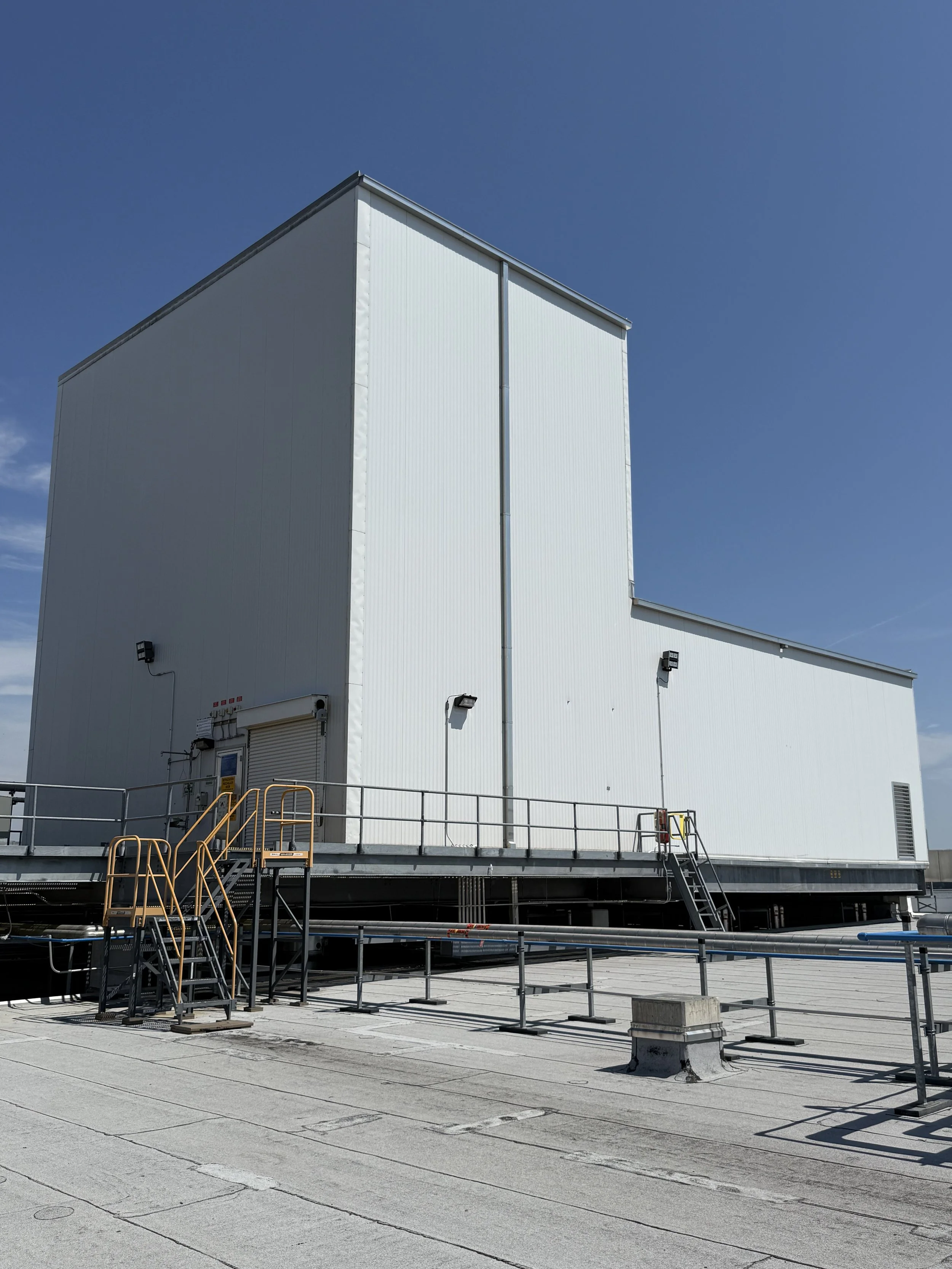 A large, white industrial building with a flat roof, set against a clear blue sky. There are exterior lights, a staircase, and a fenced platform in the foreground.