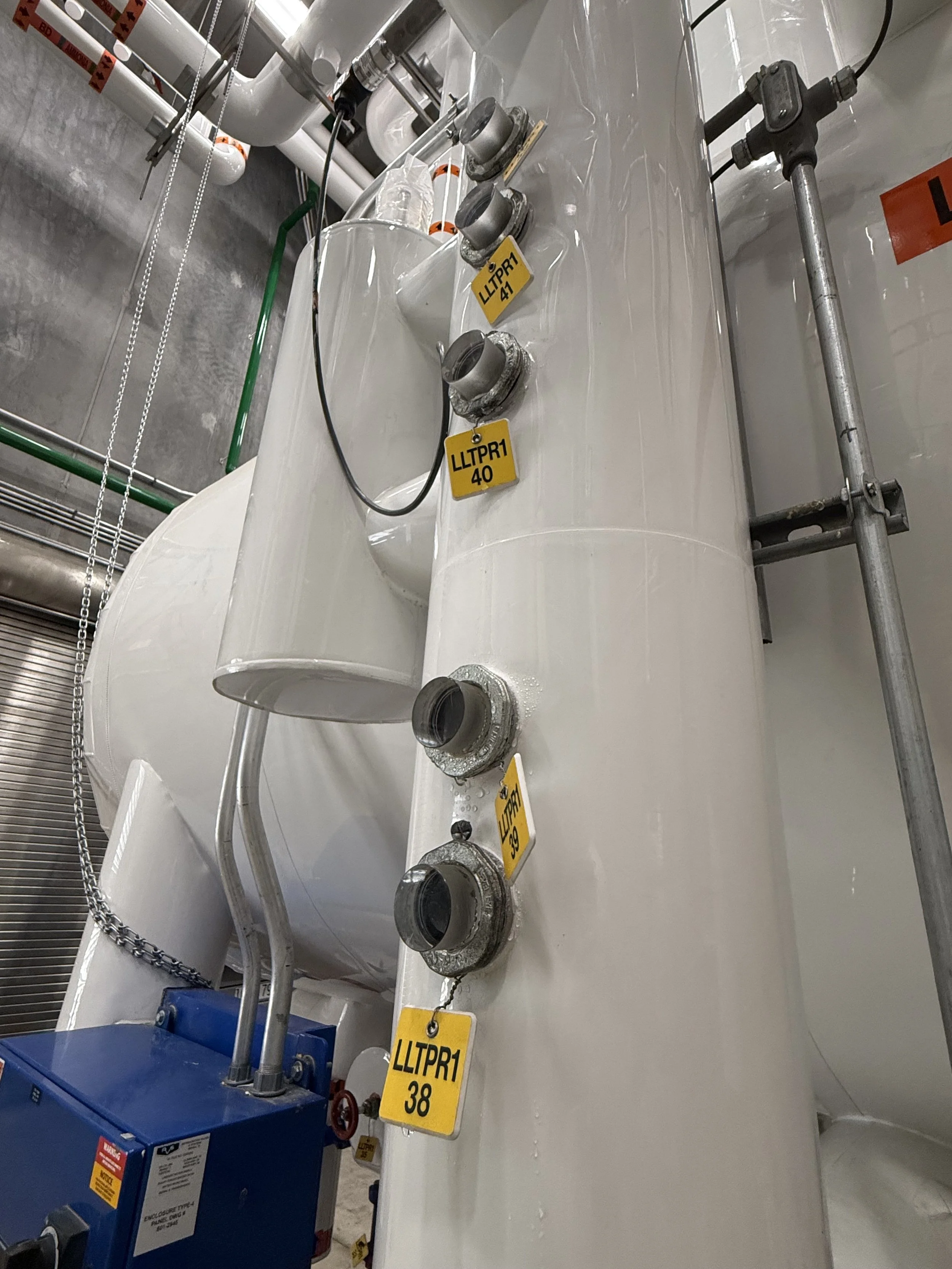 Industrial equipment with pipes, valves, and yellow tags labeled LITPR1 40, 38, and 41, connected to a large white tank or vessel, with a blue control box nearby.