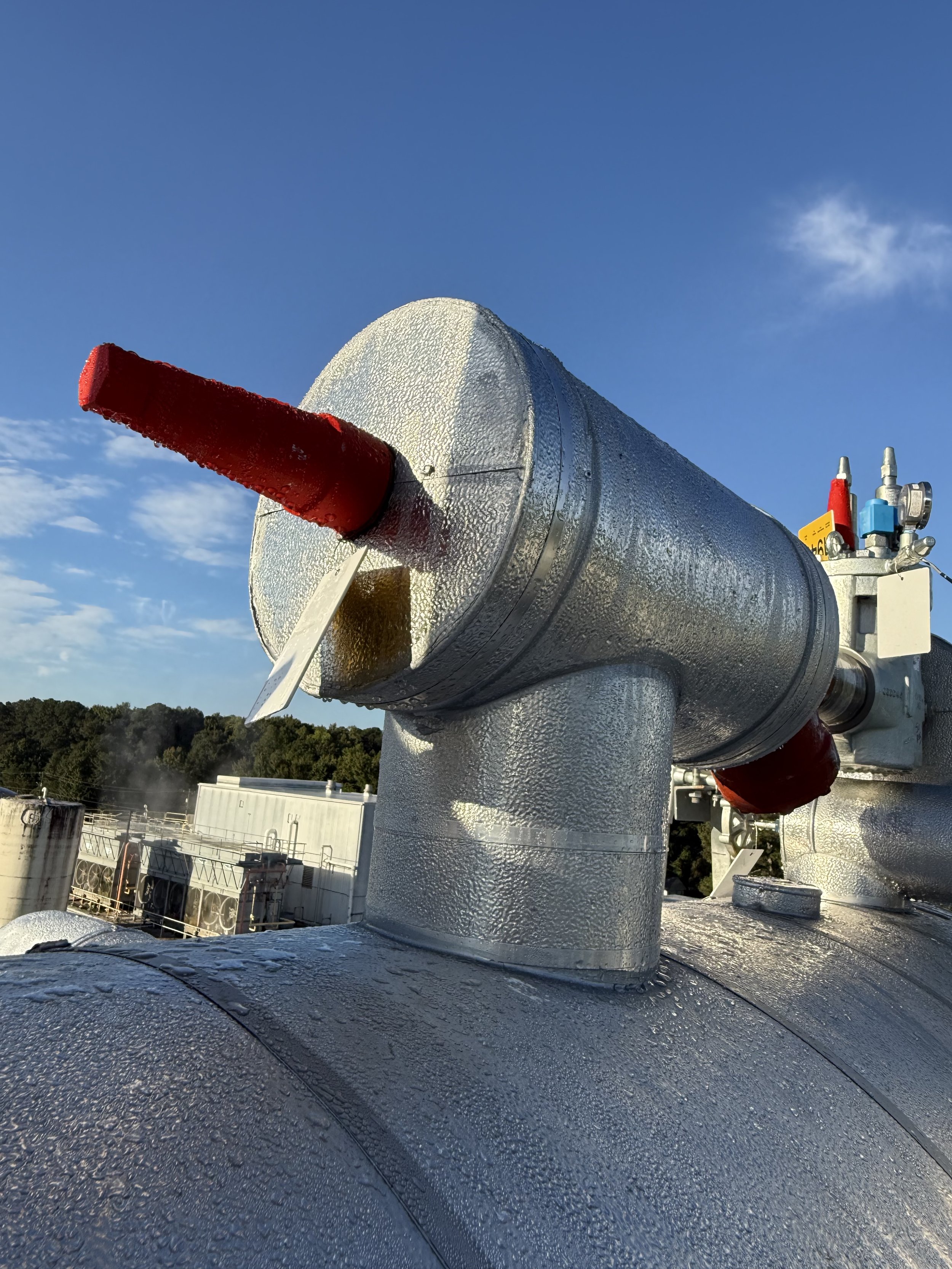Large industrial metallic pipe with red and blue valves on top, covered in water droplets, set against a clear blue sky with some clouds, and industrial buildings in the background.