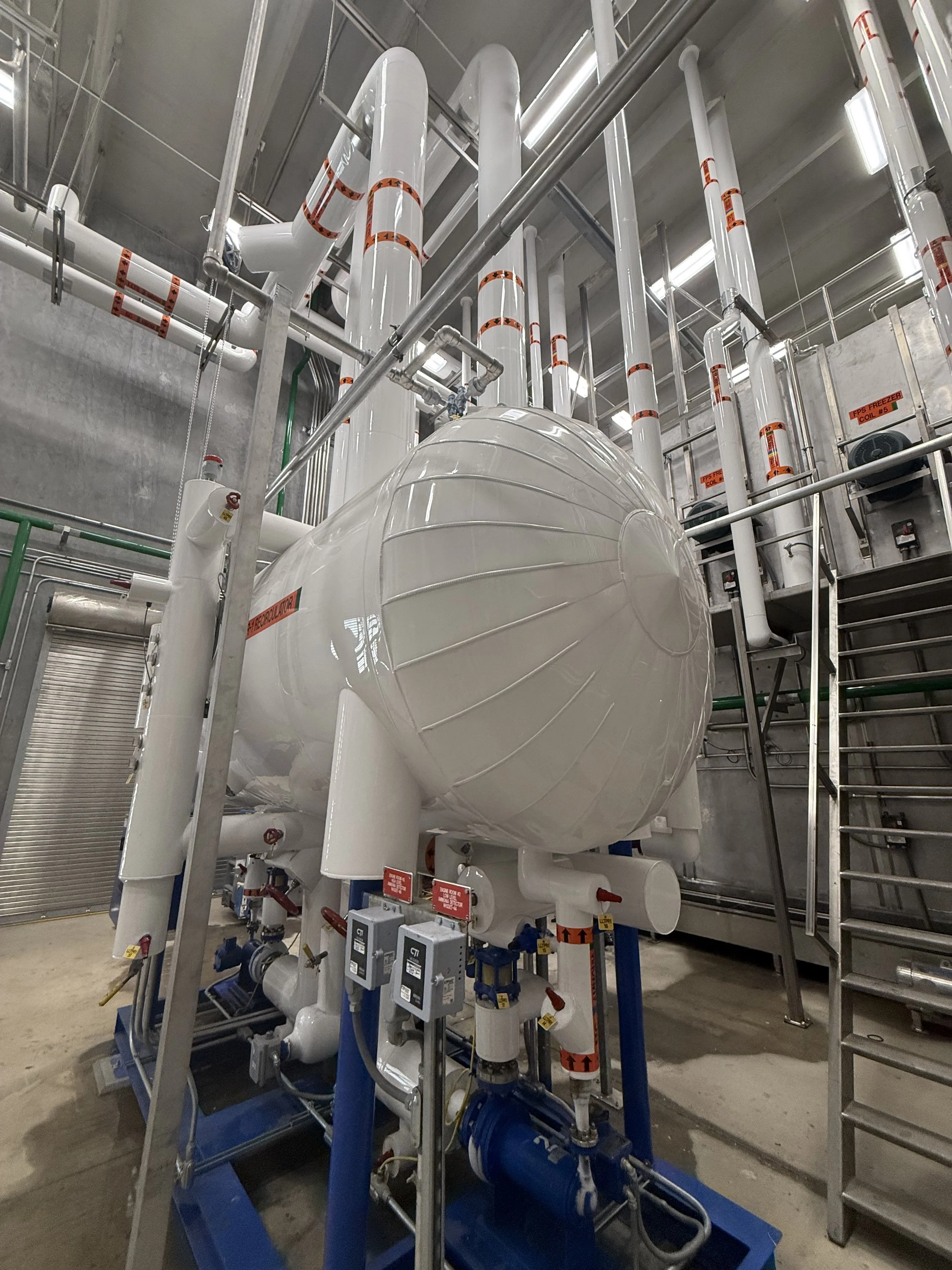 Large industrial white tank with blue piping and valves inside a building with concrete walls and ceiling, surrounded by metal scaffolding and piping.