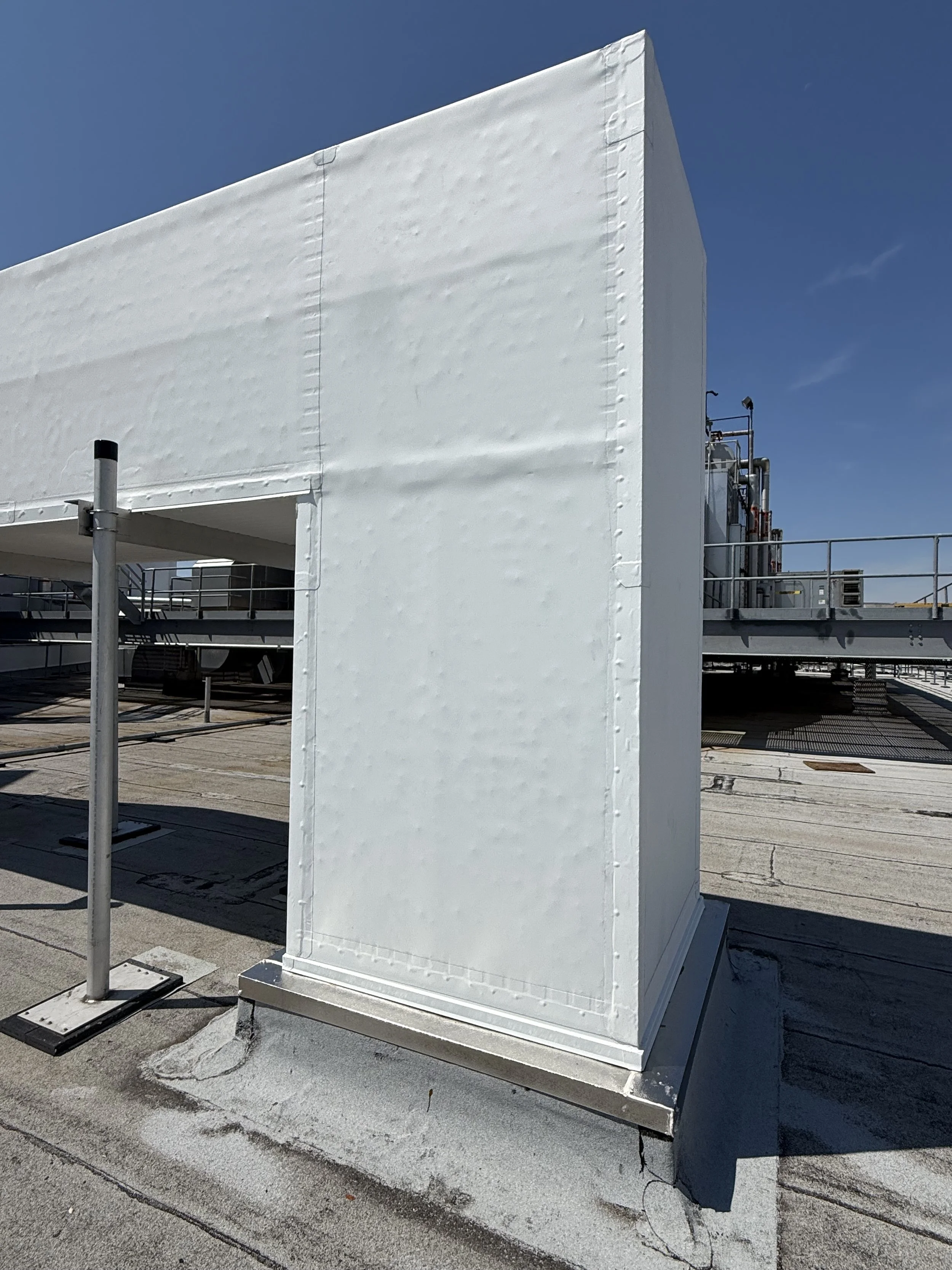 A white, enclosed rooftop structure with visible rivets on the exterior, located on a building's rooftop with a blue sky background.