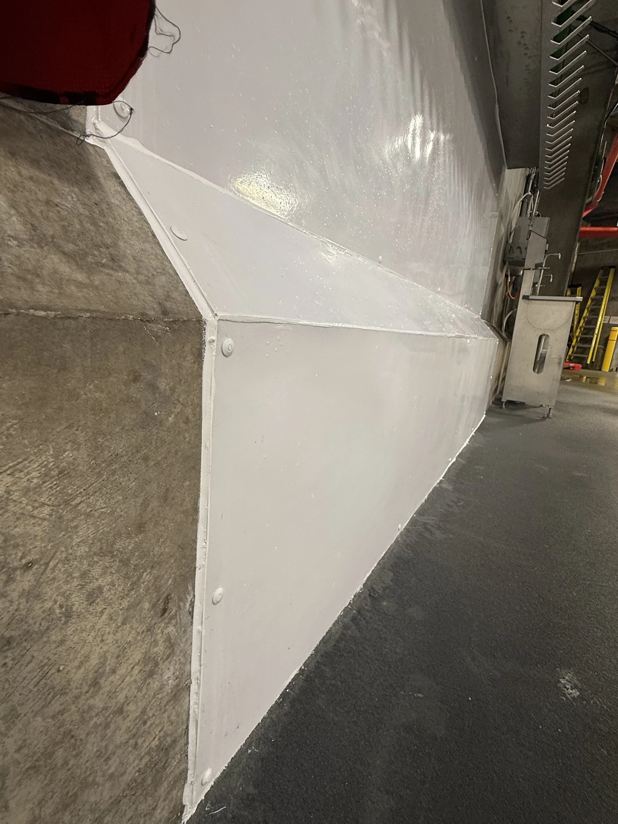 Close-up image of a corner where concrete flooring meets a white painted wall in an industrial setting with black flooring and some equipment visible in the background.