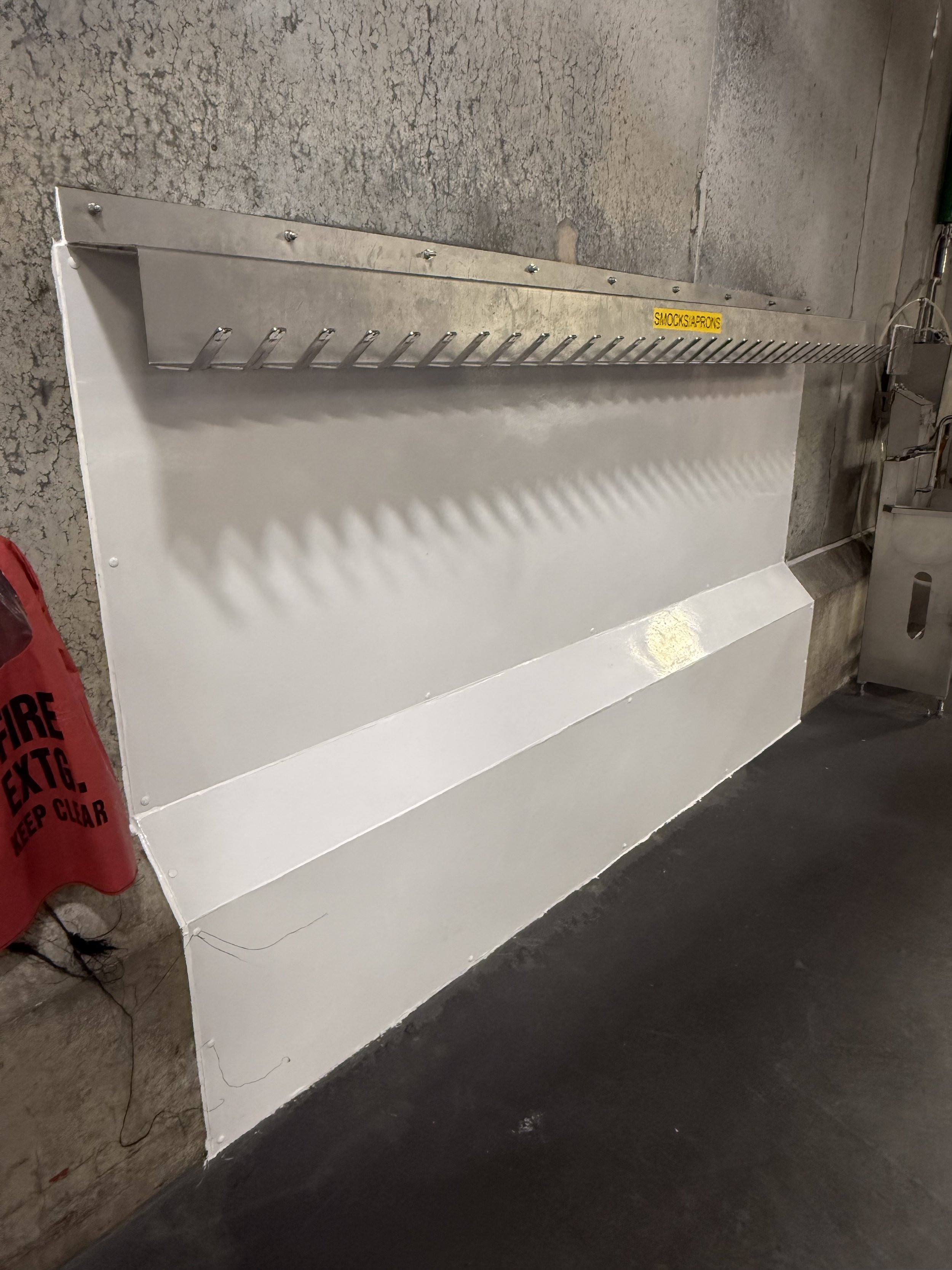 Metal pry bar or crowbar with multiple hooks resting against a white panel, mounted on a rough concrete wall. A yellow sticker with black text is attached to the metal bar, reading 'SMOCKS & APRONS'. A red fire extinguisher is partially visible on the left. Electrical cords and a device are on the right side.