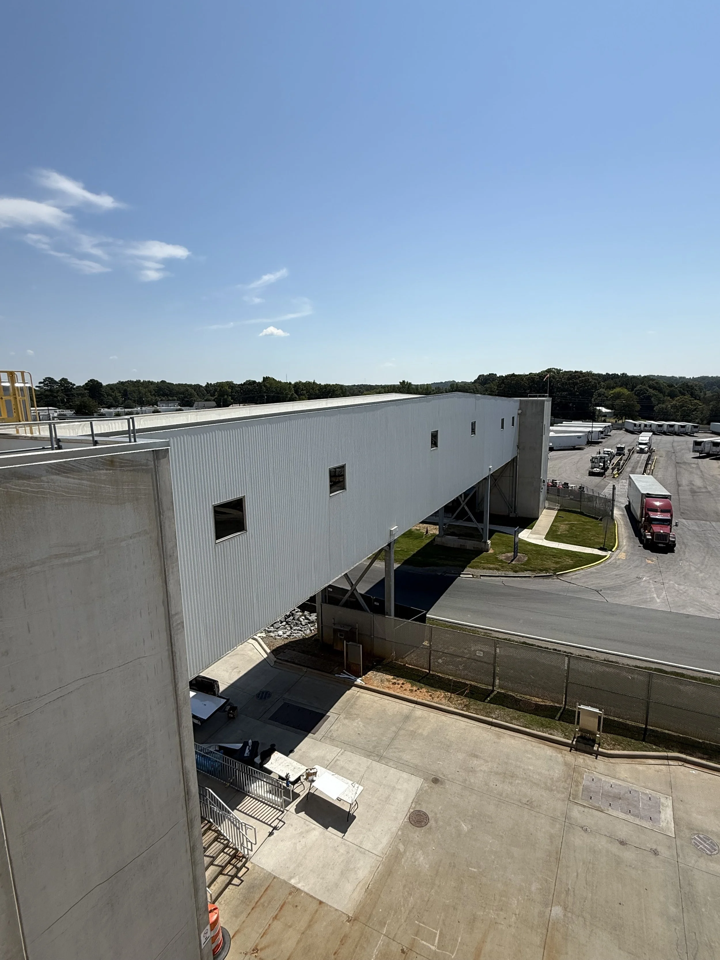 View from a high vantage point of an industrial or commercial building with a white exterior, a loading dock, and parked trucks in the background under a bright blue sky with few clouds.