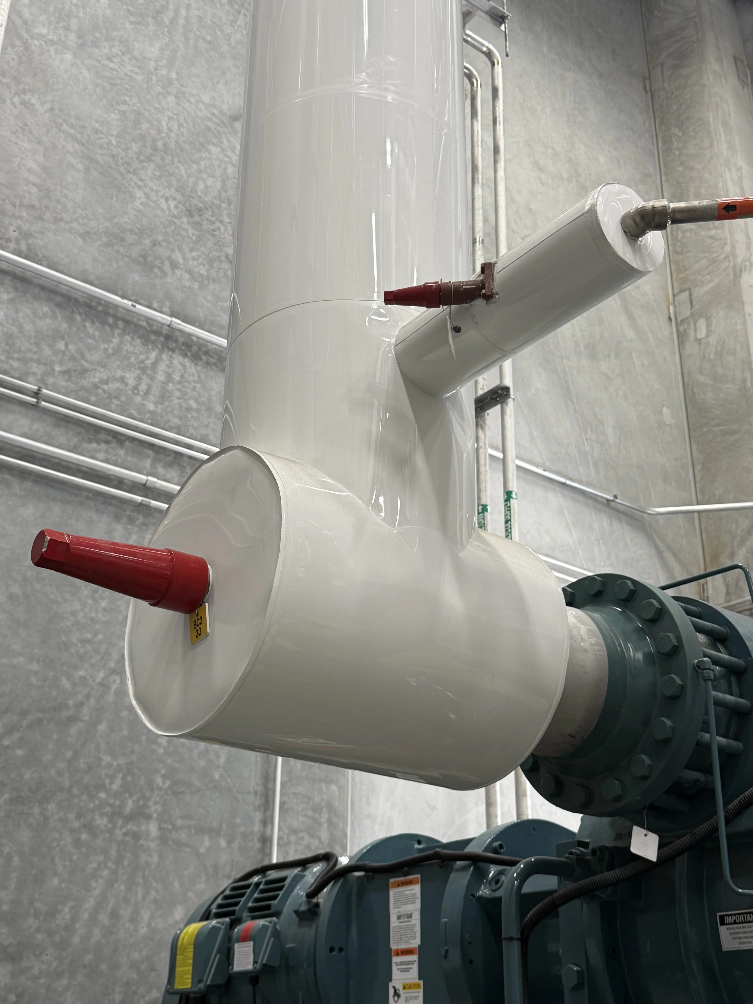 Large white industrial tank with a pipe connected to it, metal pipes and industrial equipment in the background, situated in a factory or industrial setting.