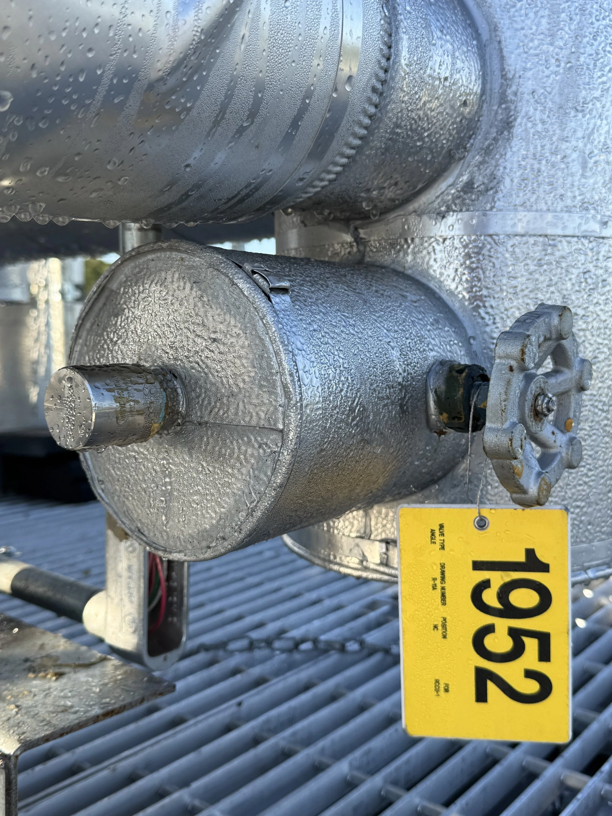 Close-up of a metallic cylindrical tank with condensation droplets on its surface, mounted on a metallic structure, with a yellow hazard tag attached containing black caution text.