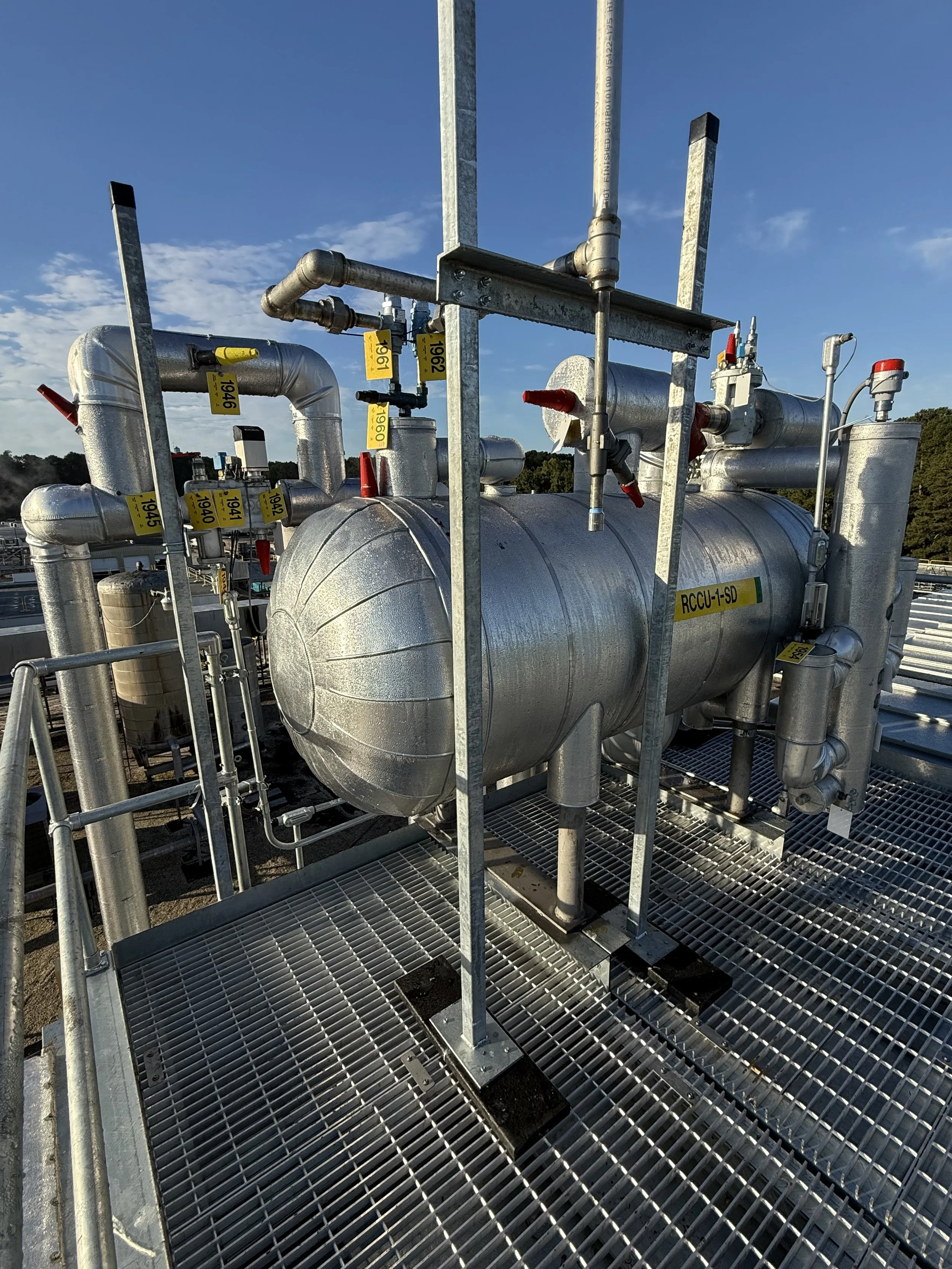 Industrial piping system with metal pipes, valves, and gauges installed on a rooftop against a blue sky with clouds.