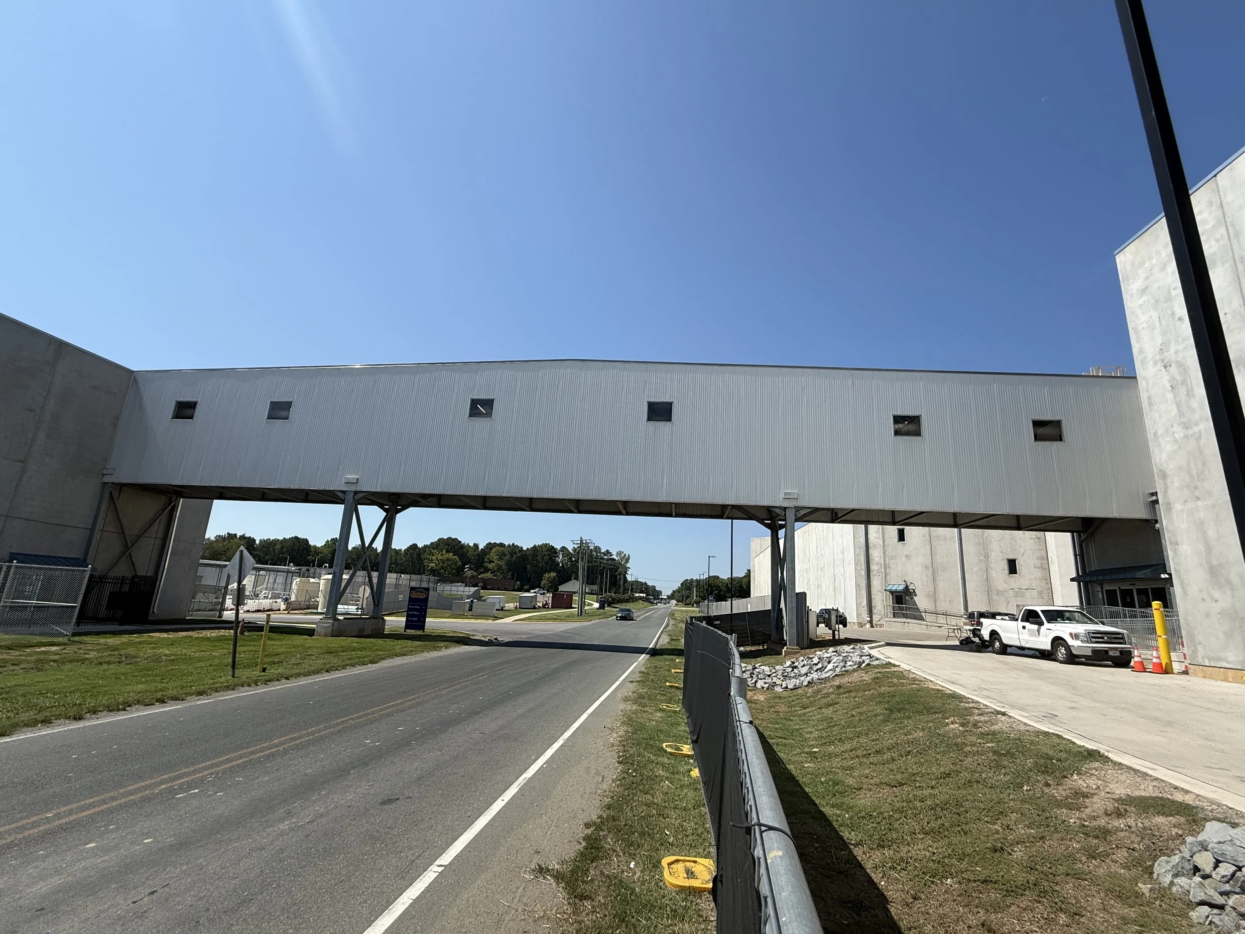 A modern building with an elevated walkway supported by metal beams, a road with cars passing underneath, and construction barriers on the sidewalk, set against a clear blue sky.