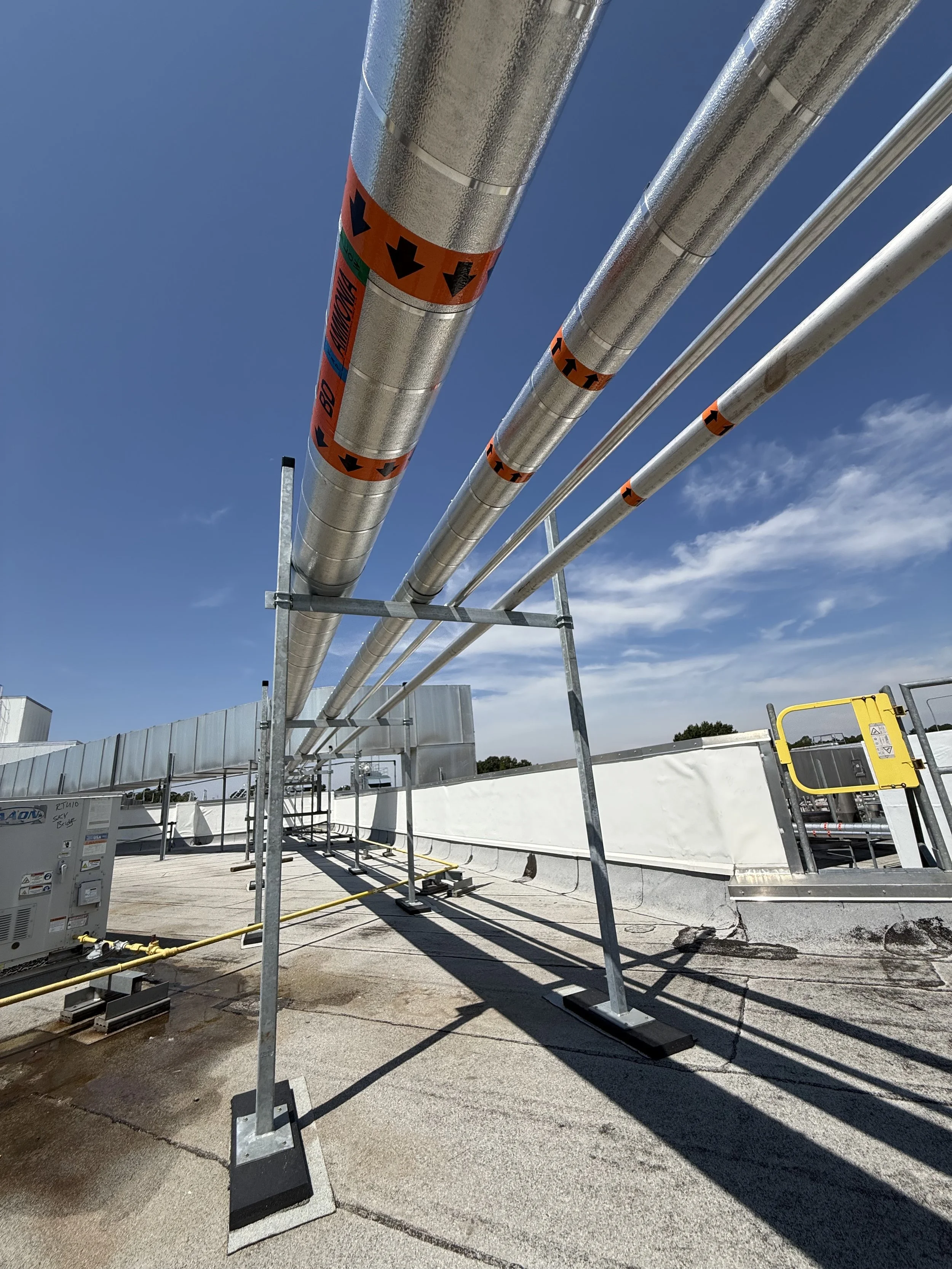 Industrial piping installation on a rooftop with large metal pipes labeled with warning stickers, supported by metal frames, under a clear blue sky.