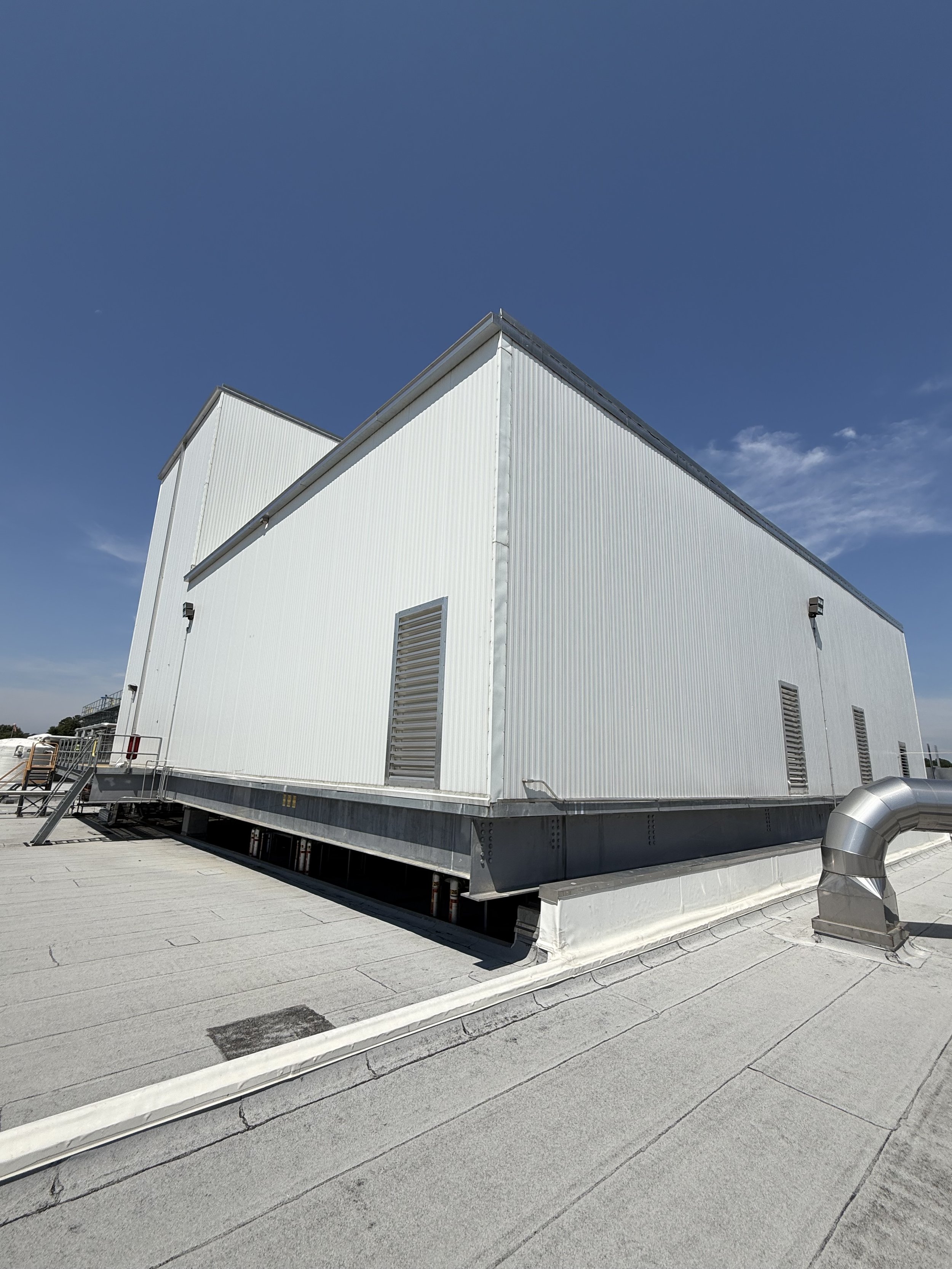 White industrial building on a flat rooftop with vents and pipes, under a clear blue sky with some light clouds.