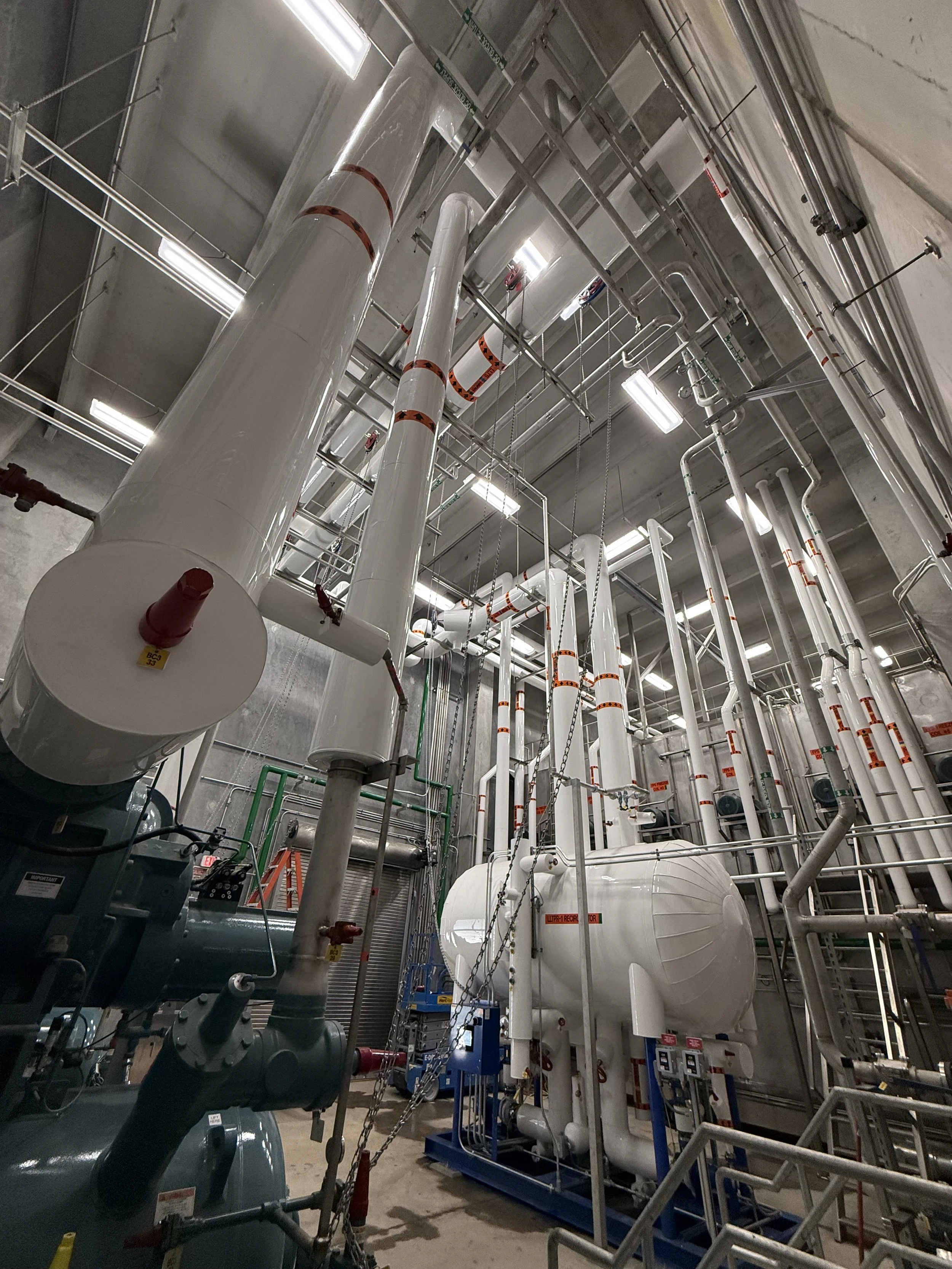 A room with large pipes and machinery, including pressure vessels and valves, indicative of an industrial or mechanical setting like a boiler room or HVAC facility.