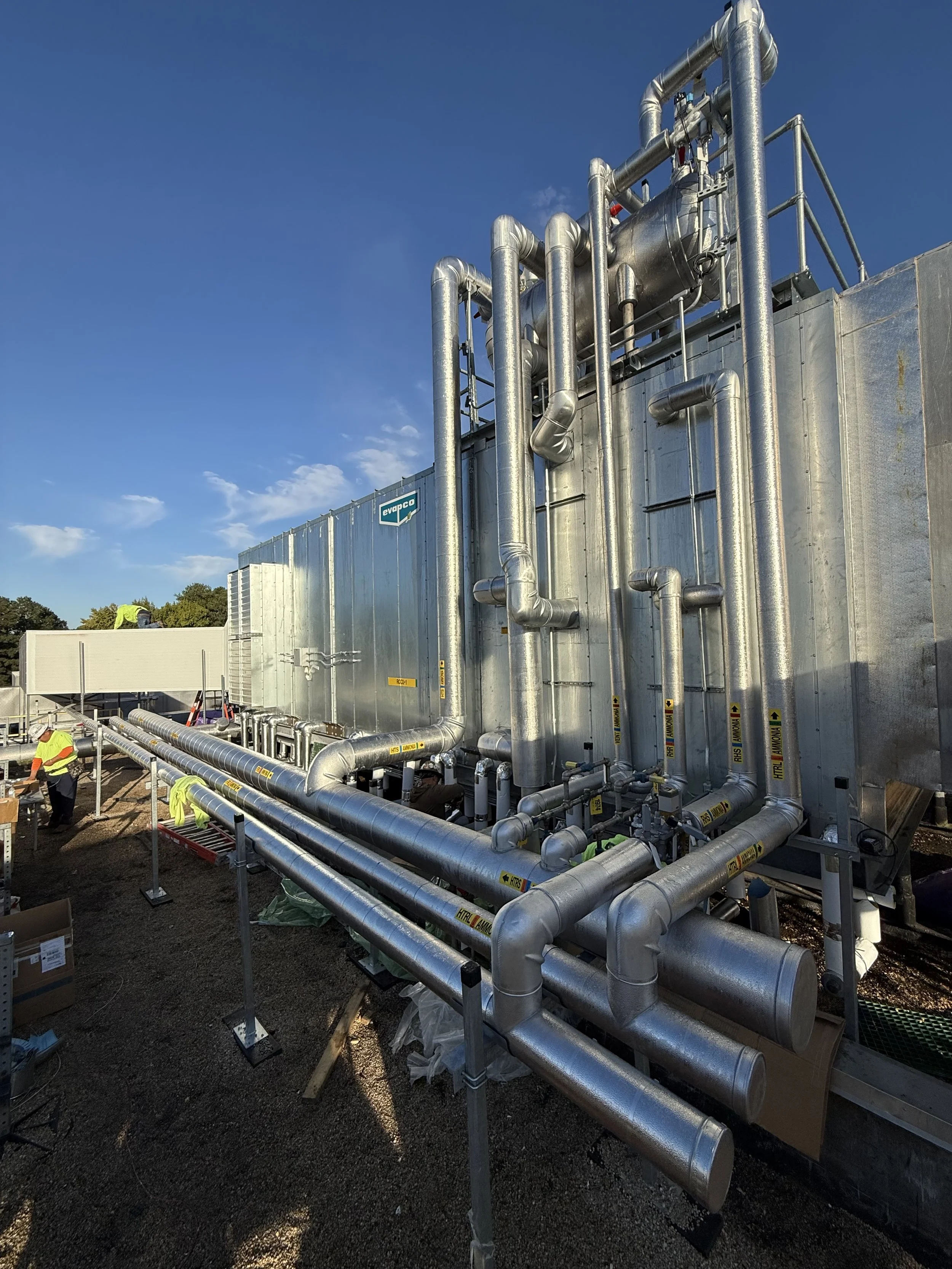Industrial rooftop with metal pipes and equipment, workers in safety gear, and a clear blue sky.