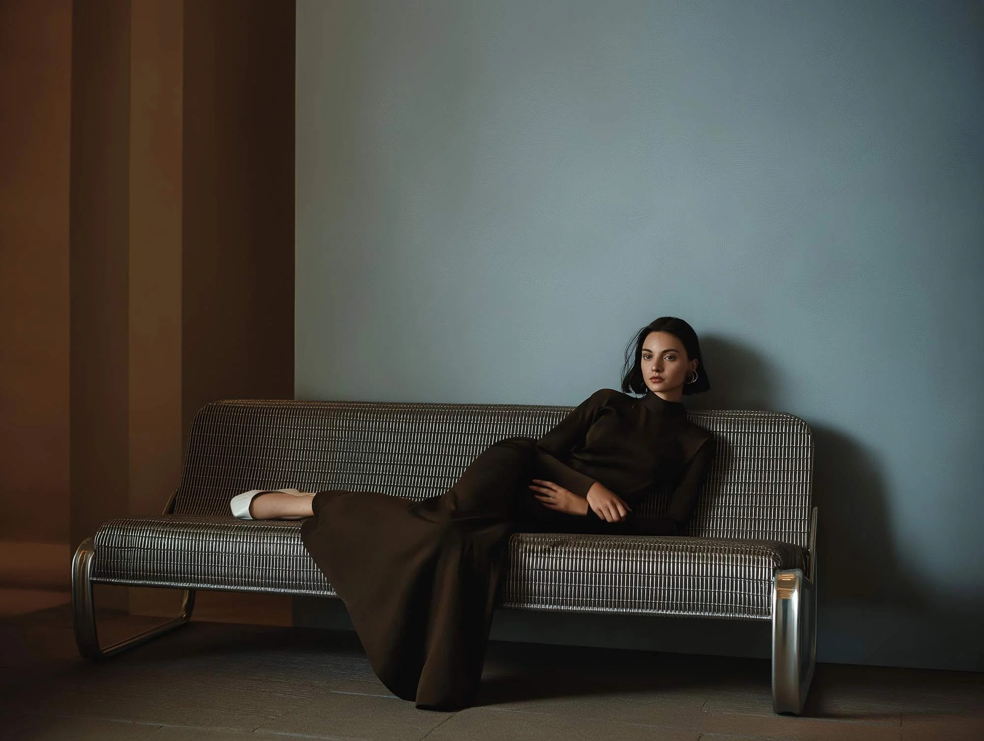 A young woman with dark hair sitting on a patterned gray sofa against a dual-colored wall, wearing a long black dress and white shoes, with a neutral expression.