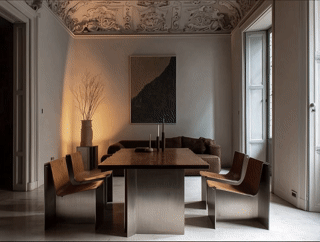 A modern dining room with a wooden table, six chairs, a vase with branches, and a large painting on the wall. Natural light filters through tall windows.