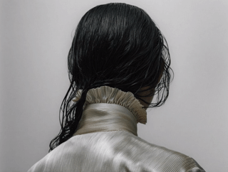 Back of person with long, wet black hair wearing a shiny, silver high-collared jacket against a plain gray background.