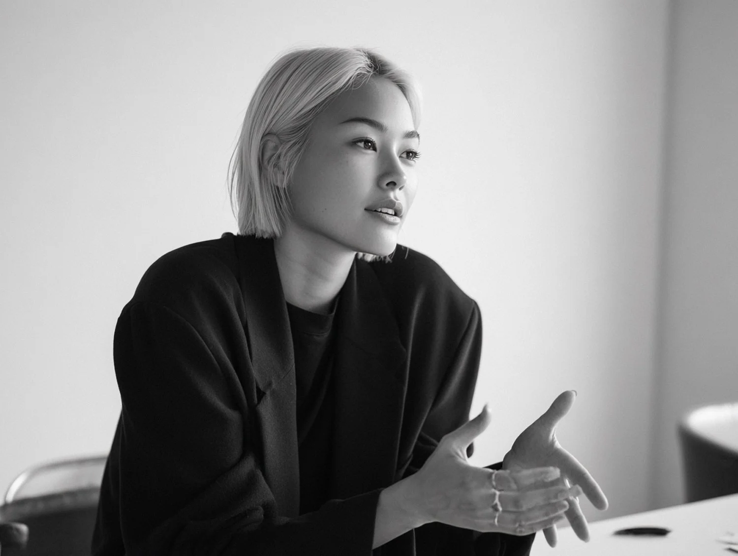 A black-and-white photo of a woman with short blonde hair, wearing a dark blazer, sitting at a table with her hands gesturing as she speaks or explains something, in a minimalistic setting.