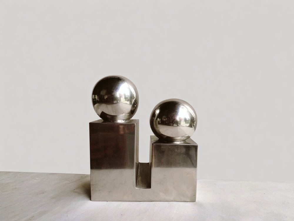 Modern metallic sculpture with two shiny silver spheres on different height rectangular bases, set against a plain wall background.