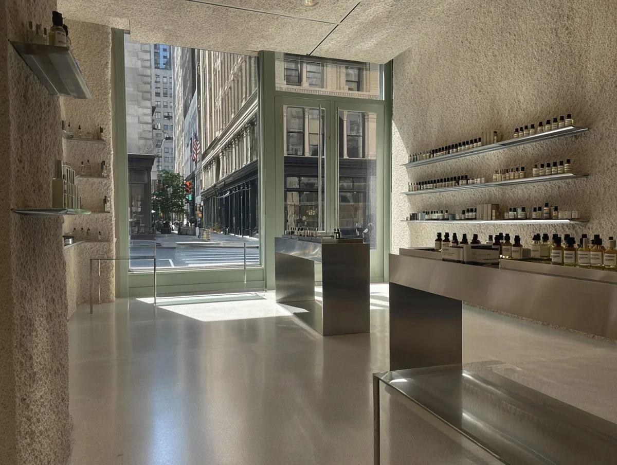 A minimalist store interior with textured beige walls, white flooring, and shelves with small bottles, sunlight streaming through large glass doors overlooking a city street.
