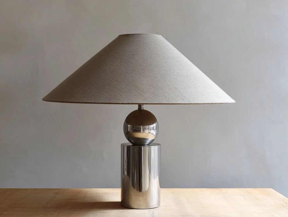 A modern table lamp with a metallic base, spherical accent, and beige fabric shade, placed on a wooden surface against a plain wall.