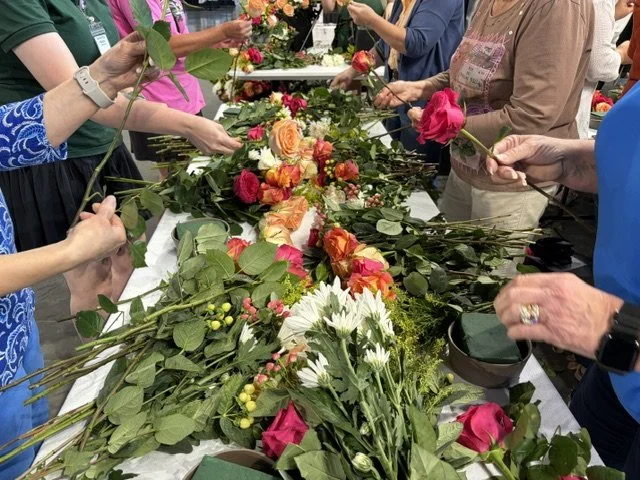 People arranging and wrapping colorful flowers, including roses and daisies, on a long table.