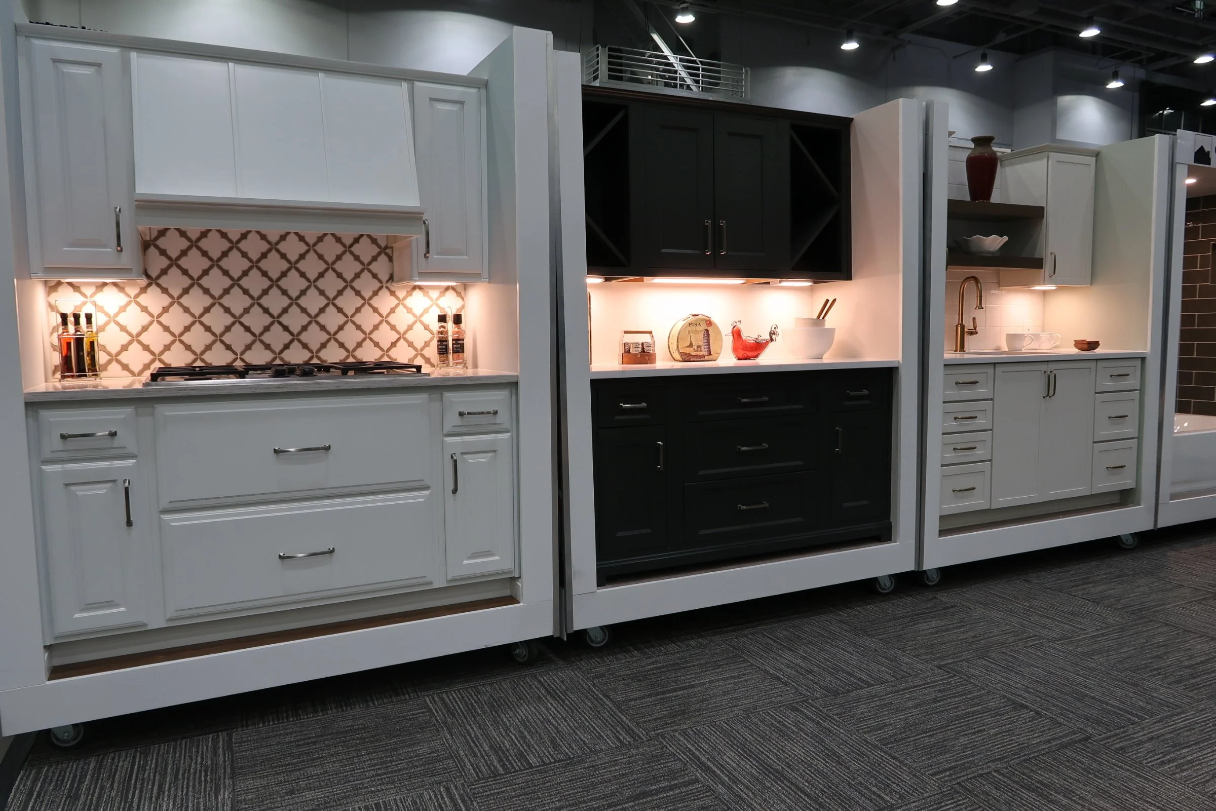 Display of kitchen cabinets in white and black with under-cabinet lighting, decorated with kitchenware and decorative items, set on a showroom carpet.