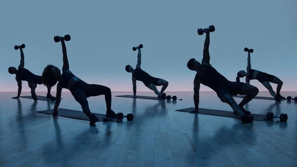 Group of people exercising with dumbbells on yoga mats in a fitness studio at sunset.