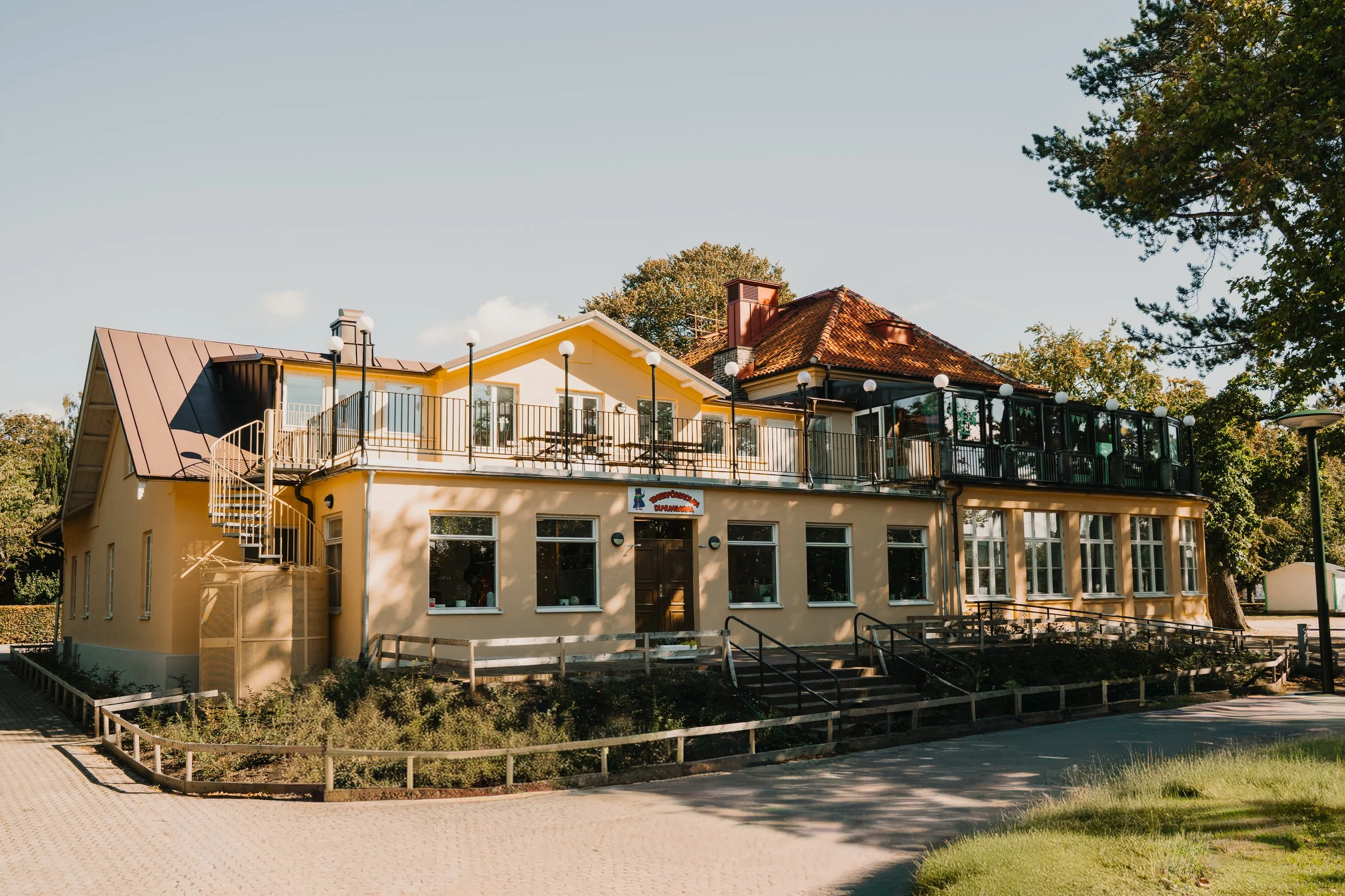 En tvåvåningsbyggnad med en uteservering på taket, omgiven av träd och ett litet räcken. Gula och vita väggar, stora fönster, och ett spirande trapphus på sidan.