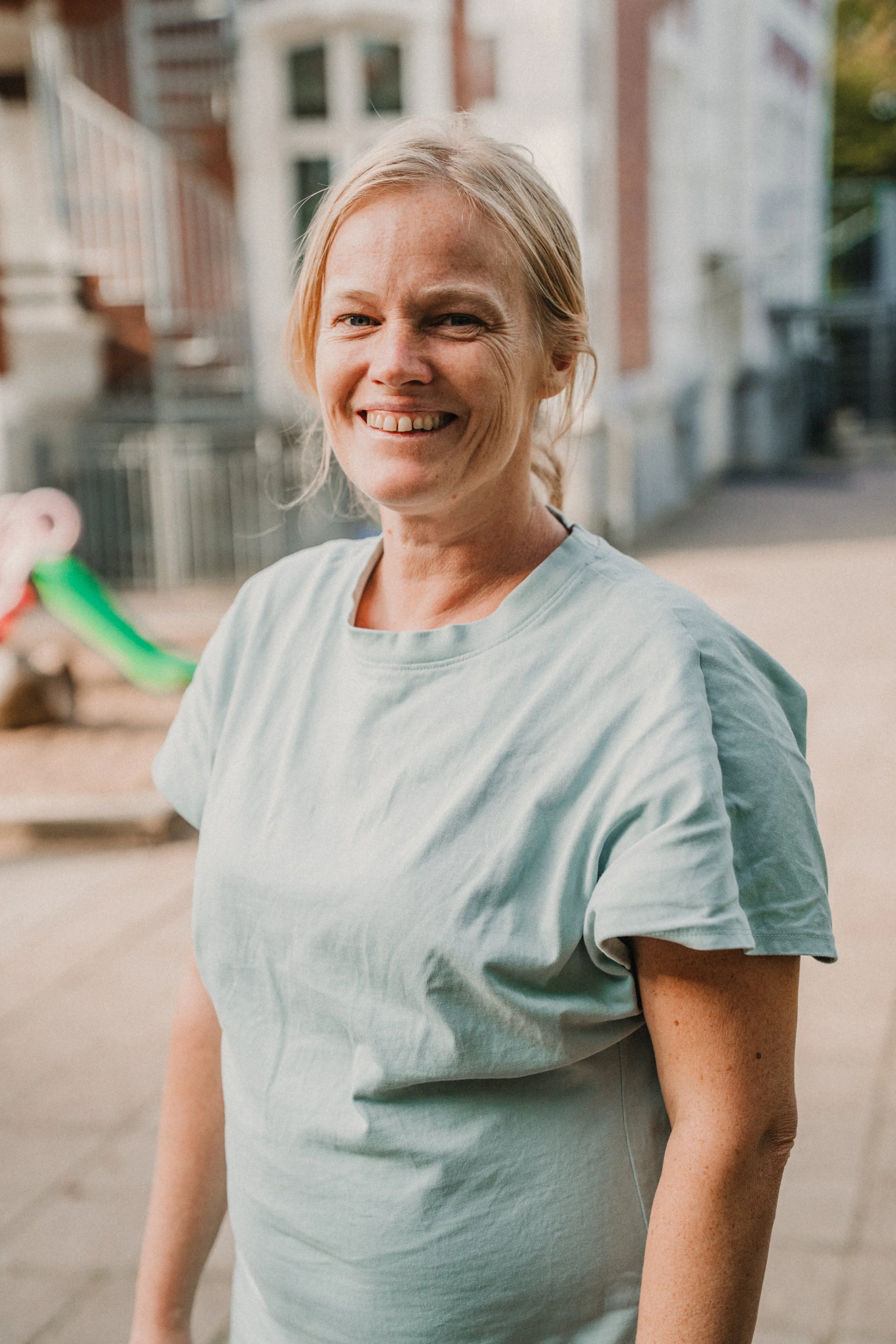 Glad kvinna med ljus hår, iklädd ljusblå T-shirt, står utomhus i ett bostadsområde med byggnader och lekplats i bakgrunden.