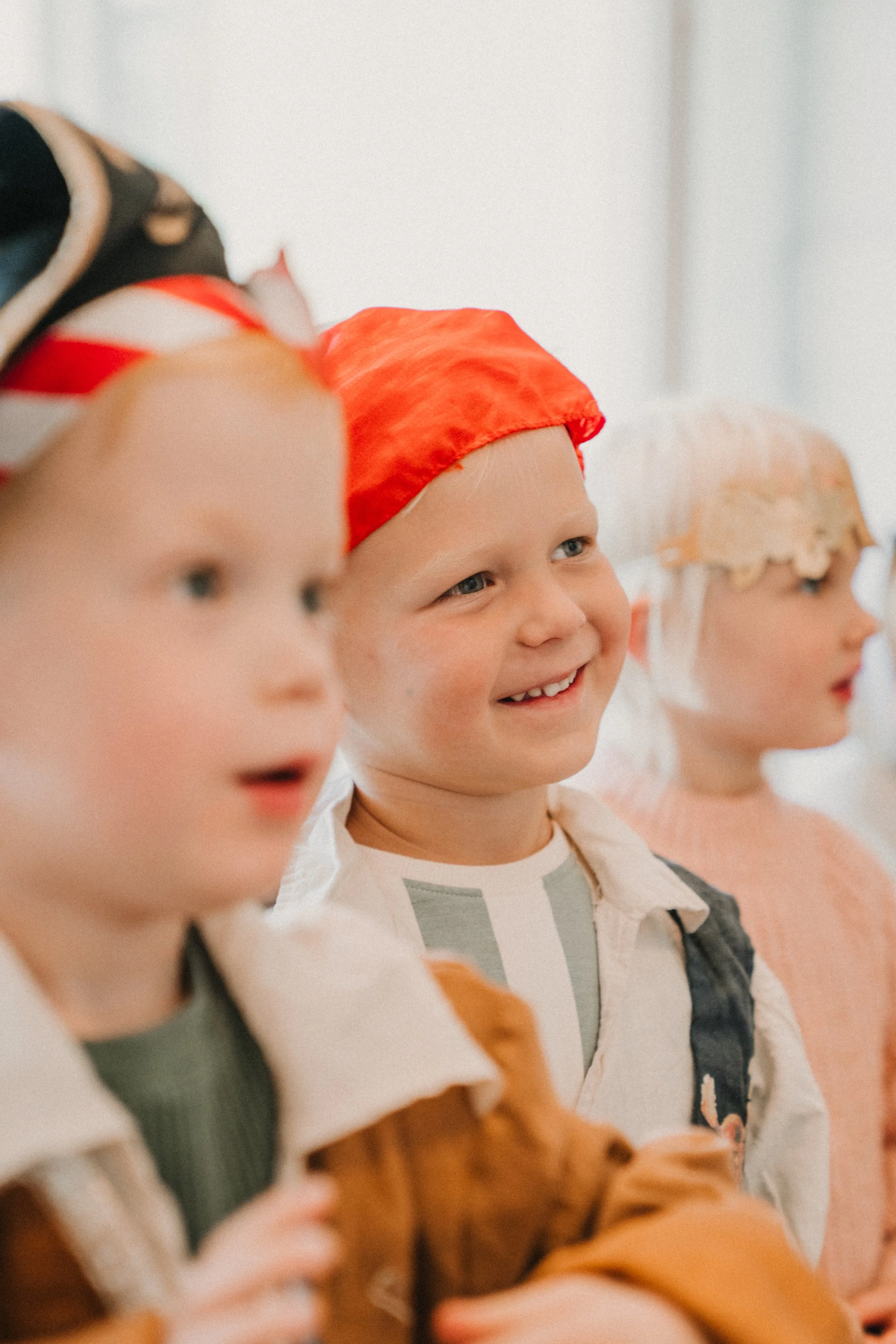 Grupp av barn i förskoleålder som tittar och ler, klädda i färgglada kläder och mössor, i ett inomhusmiljö.