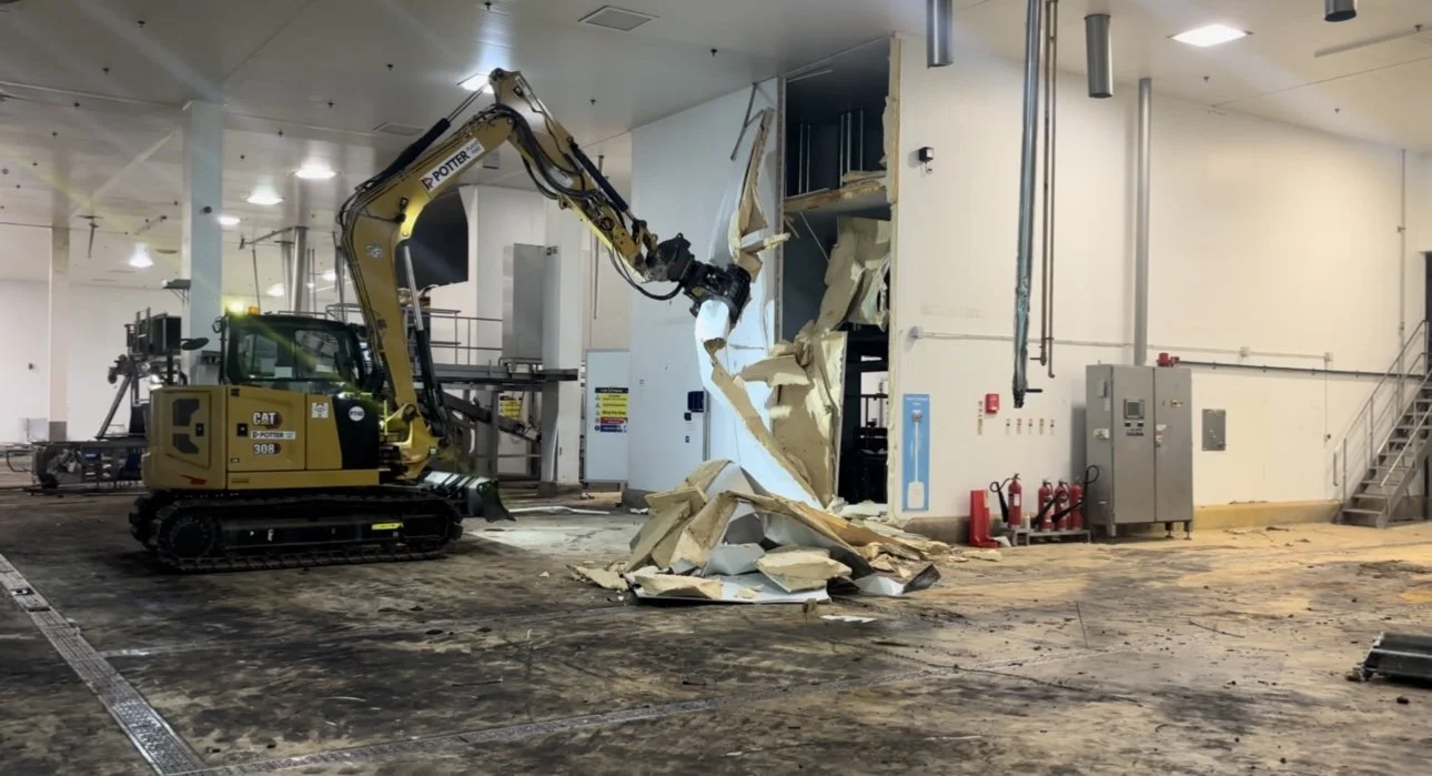 A construction excavator demolishing a wall inside a building, with debris on the floor.