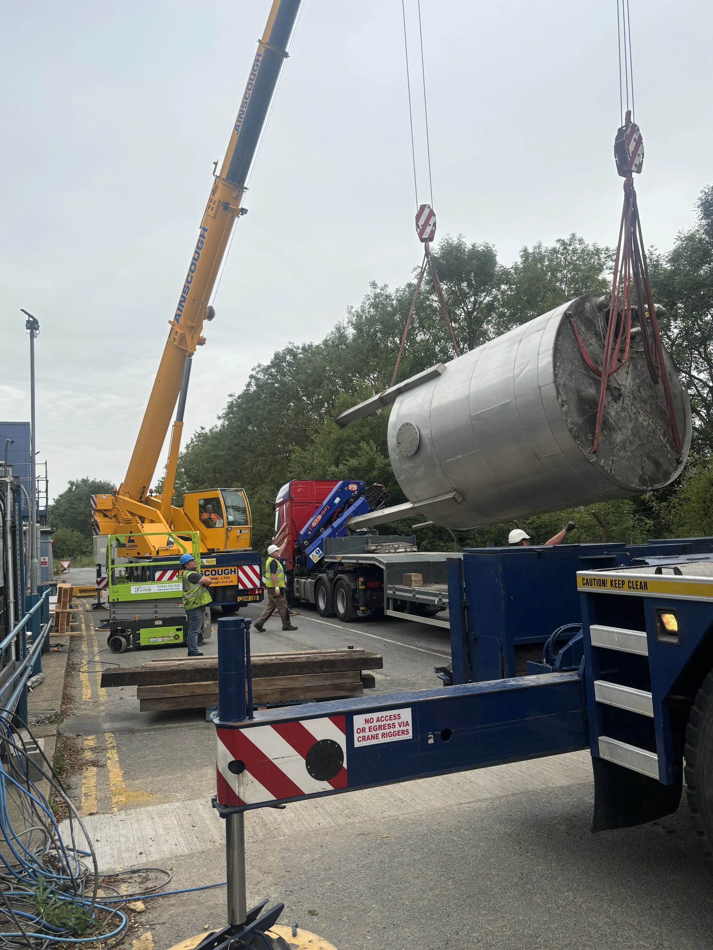 Workers guiding a large stainless steel tank being lifted by a crane at a construction site, with signage indicating restricted access.