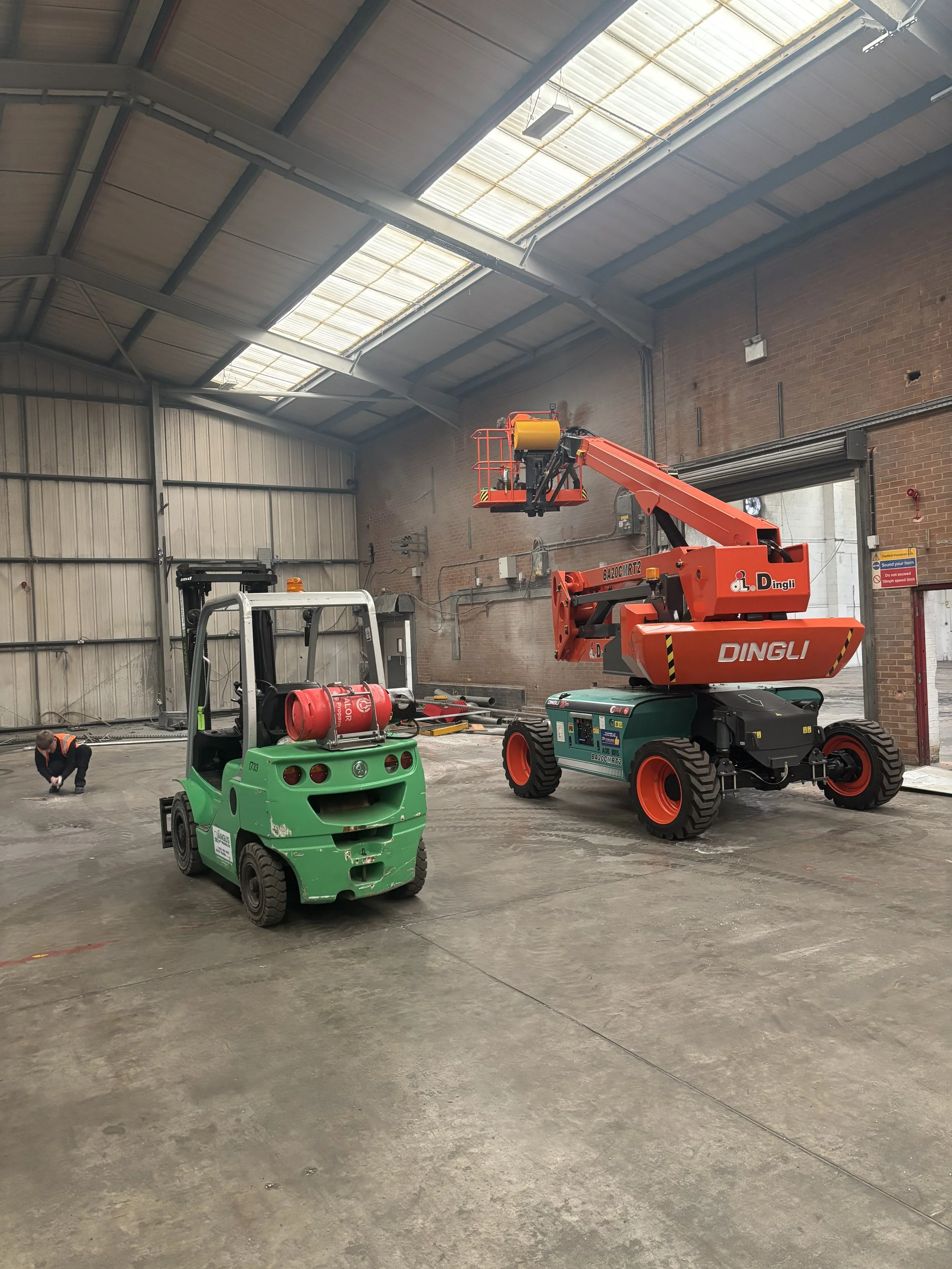 Inside a large industrial warehouse with a high ceiling, featuring skylights. There is a green forklift with a fire extinguisher mounted on it and a red telescopic boom lift. A worker is kneeling on the concrete floor, possibly inspecting or working.