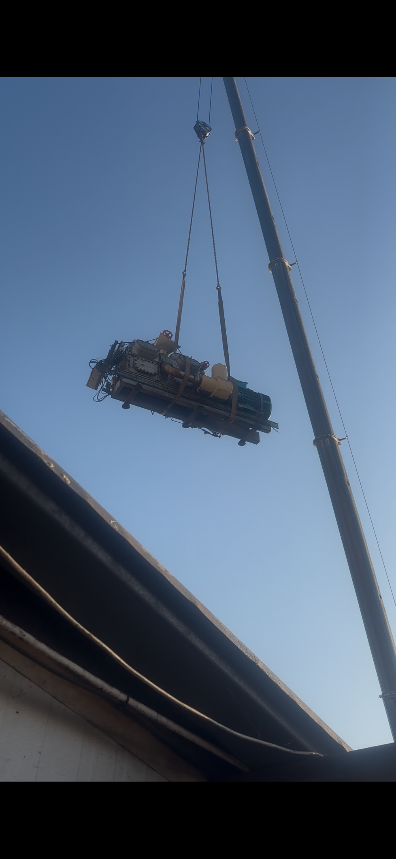 A large piece refrigeration compressor, is being lifted by a crane against a clear blue sky.