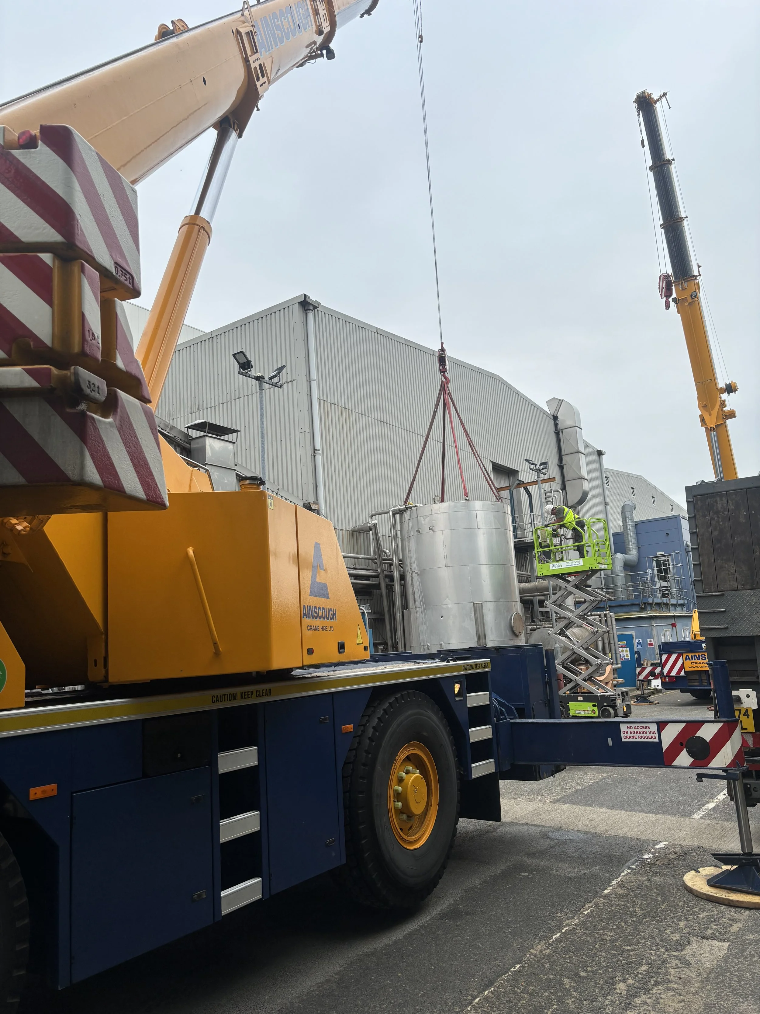 Construction scene with a yellow crane lifting a large cylindrical metal tank, workers in safety vests and helmets guiding the process, and a construction vehicle with a built-in elevated work platform.