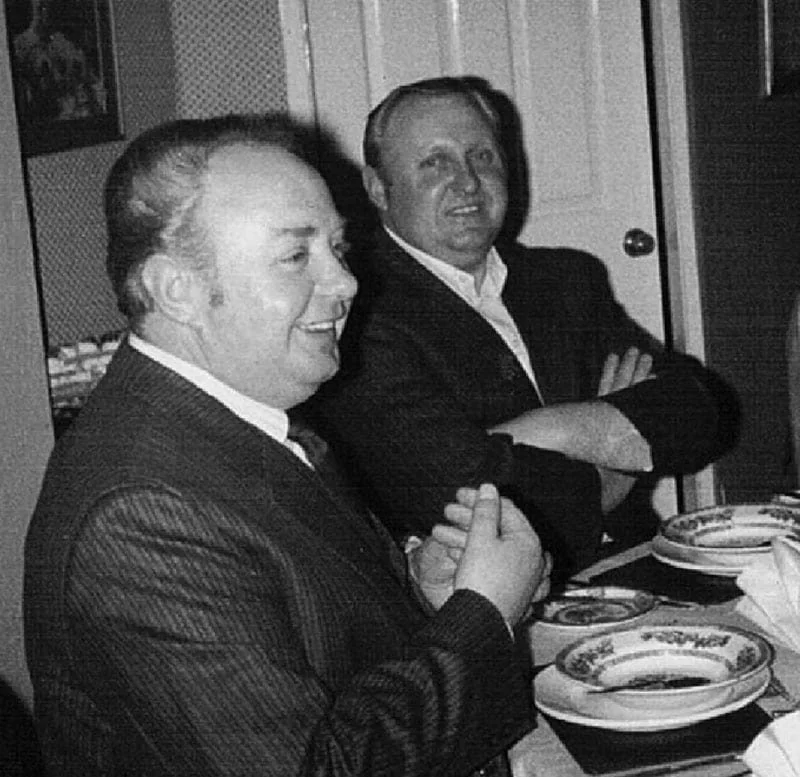 Two men in suits sitting at a dining table, smiling and facing the camera in a black-and-white photo david and richard sandham.