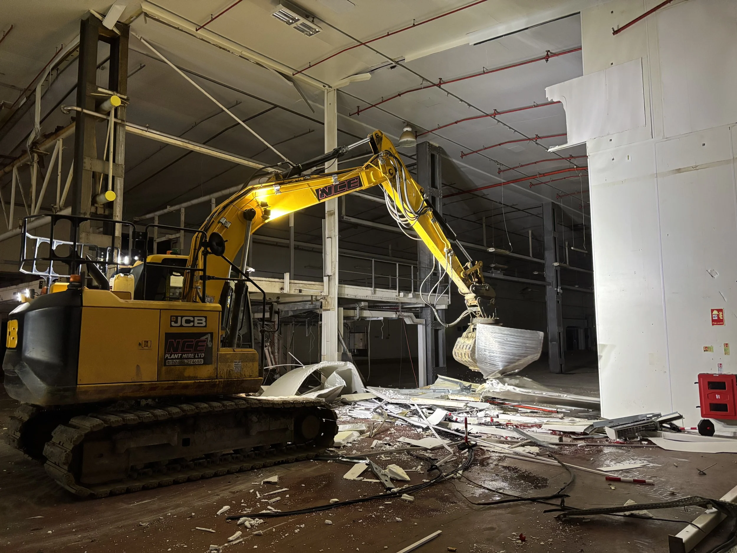 Construction or demolition site inside a building with debris scattered on the floor. A large yellow excavator with the brand JCB is positioned on the left side, lifting a large piece of debris. The ceiling has exposed pipes and ducts, and the walls are partially unfinished or damaged.