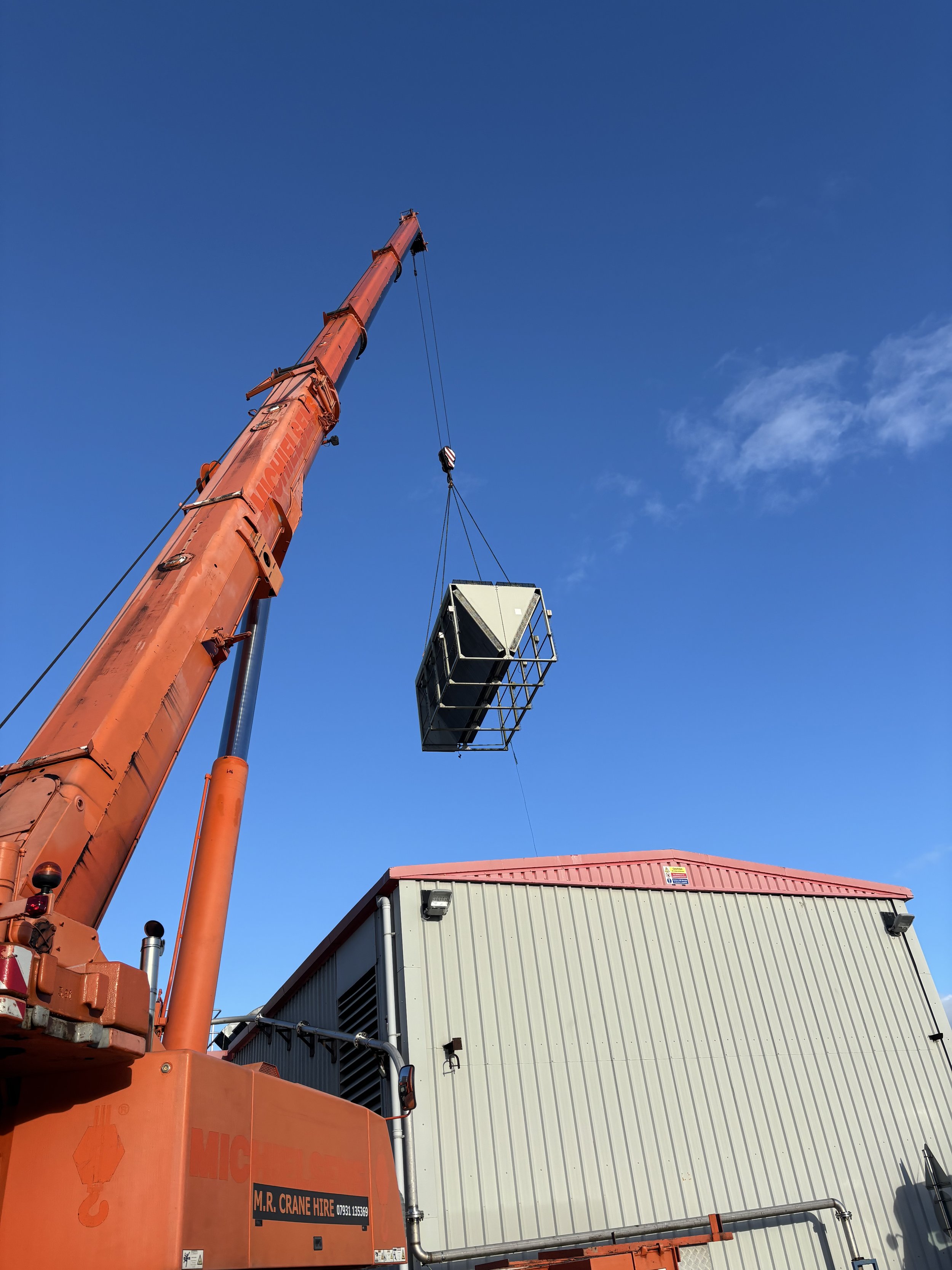 A large orange crane lifting a water chiller in front of a gray building with a red roof and a blue sky with some clouds.