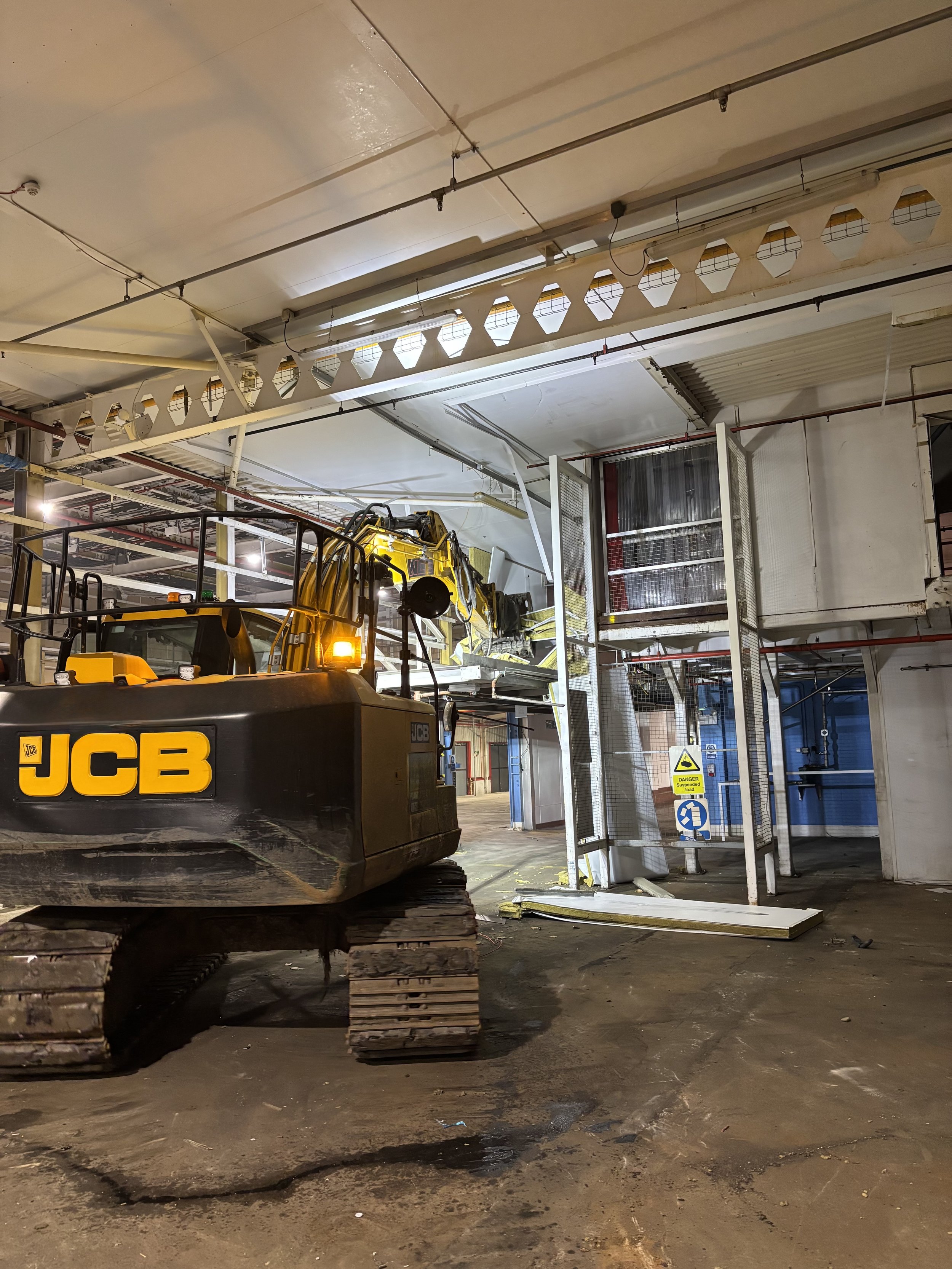 Construction site with heavy machinery, including a yellow JCB excavator, inside an industrial building with exposed ceiling pipes, metallic structures, and a partially dismantled wall with signage.